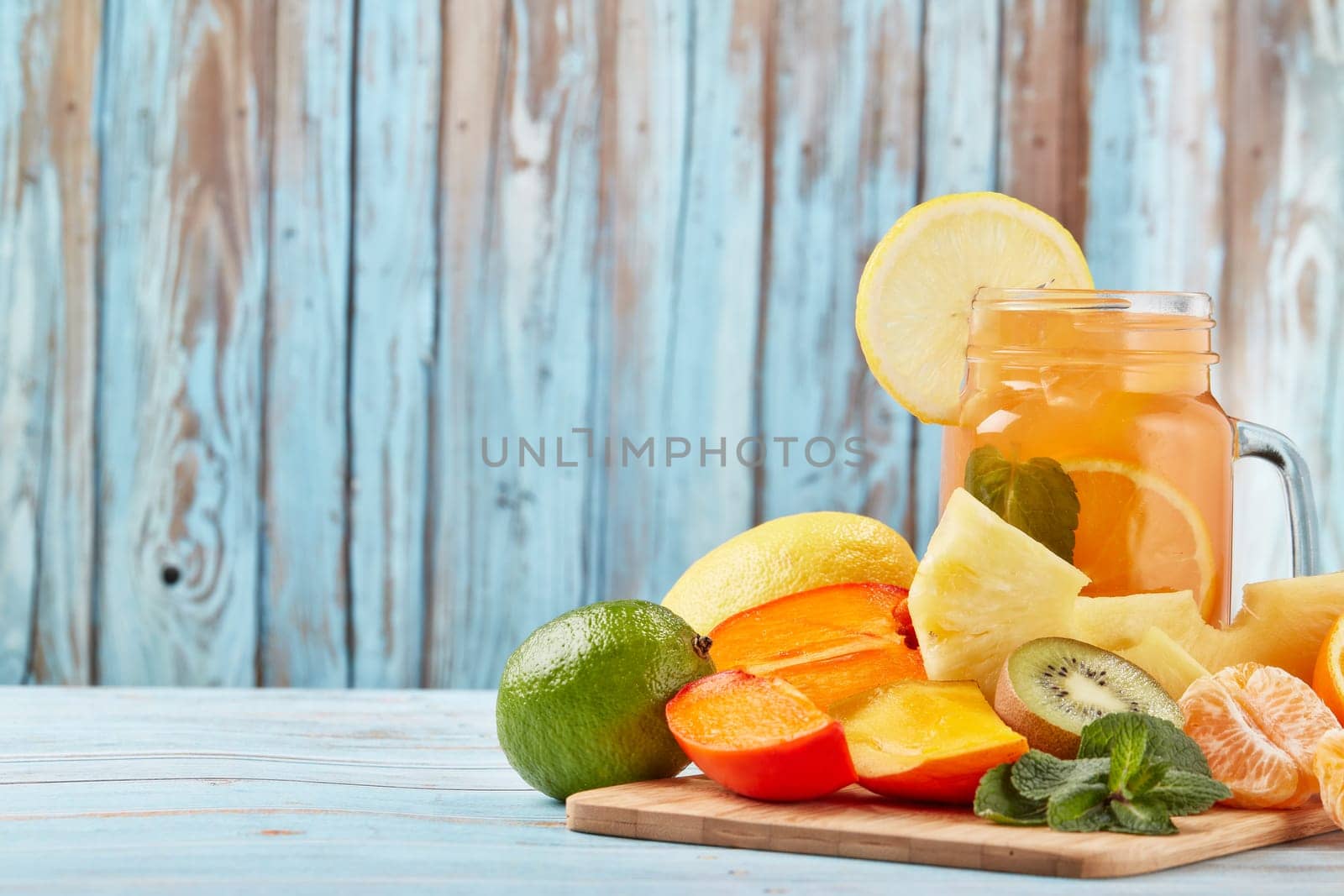 Fruit lemonade with ice, leaves of mint, lemon, pineapple, mango and lime slices standing on blue wooden table. Homemade lemonade with mix fruits. Healthy cool beverage, fruit juice