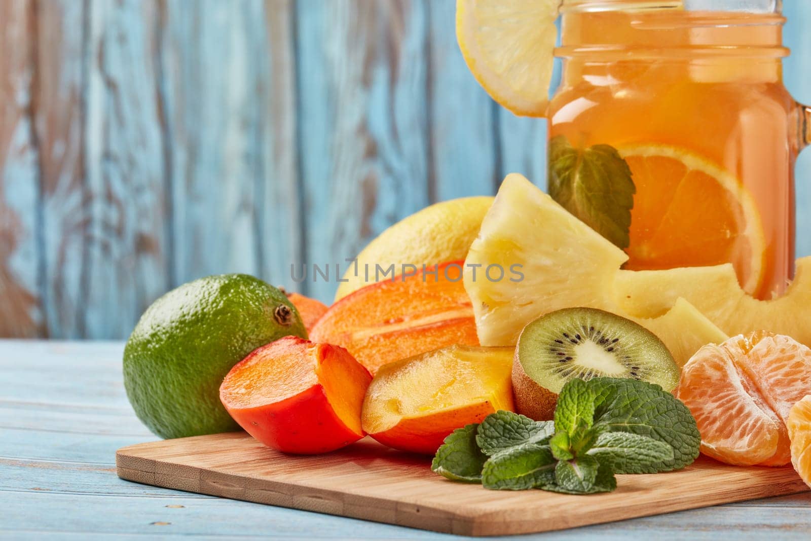Fruit lemonade with ice, leaves of mint, lemon, pineapple, mango and lime slices standing on blue wooden table. Homemade lemonade with mix fruits. Healthy cool beverage, fruit juice