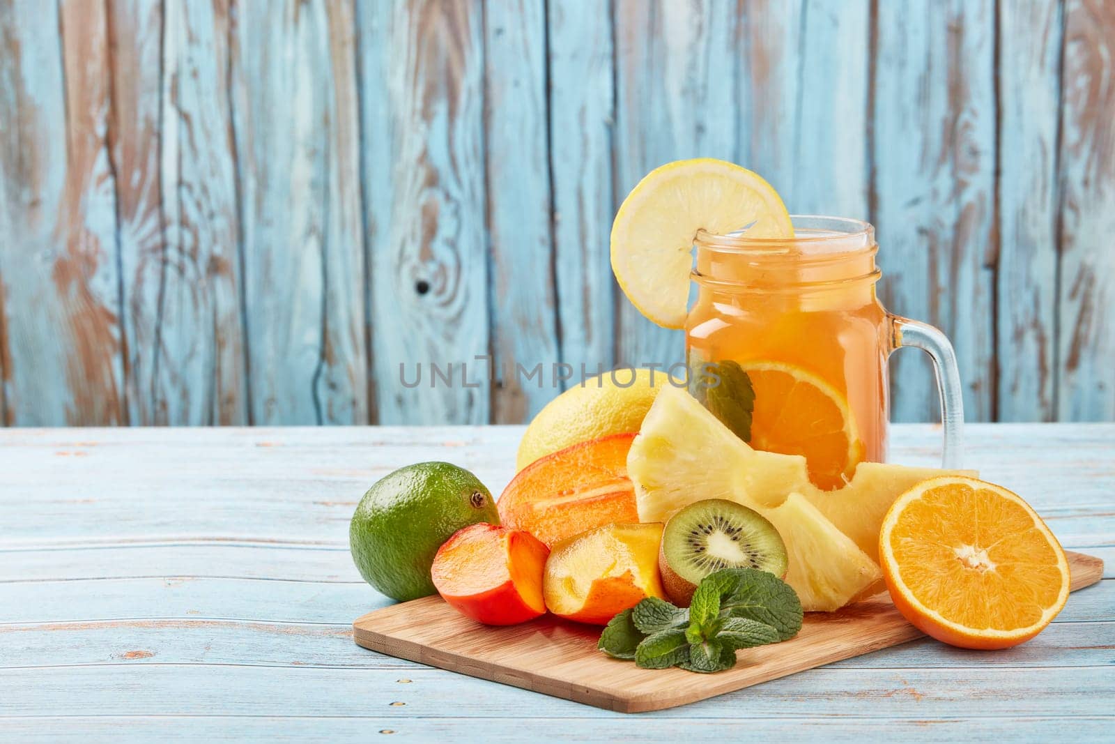 Fruit lemonade with ice, leaves of mint, lemon, pineapple, mango and lime slices standing on blue wooden table. Homemade lemonade with mix fruits. Healthy cool beverage, fruit juice