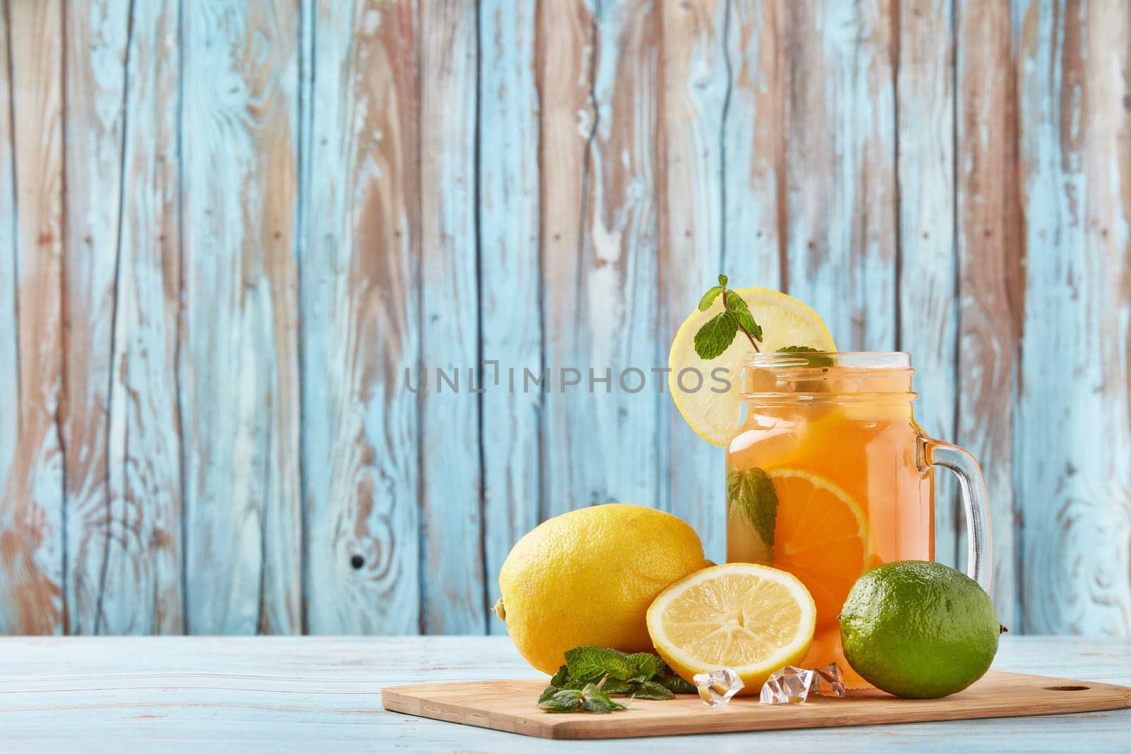 Citrus lemonade with ice, leaves of mint, lemon and lime slices standing on blue wooden table. Homemade lemonade with citruses. Healthy cool beverage