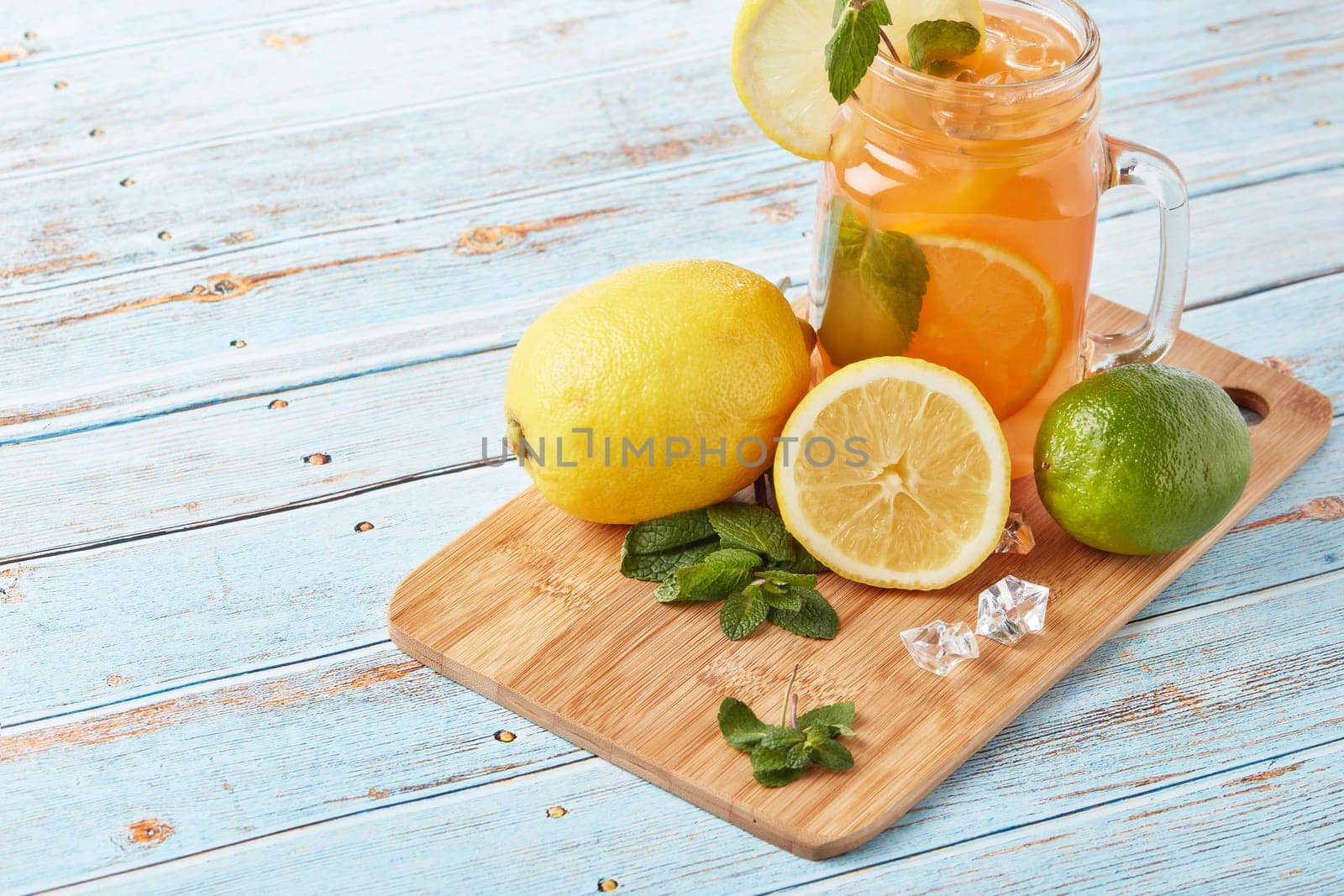 Citrus lemonade with ice, leaves of mint, lemon and lime slices standing on blue wooden table. Homemade lemonade with citruses. Healthy cool beverage