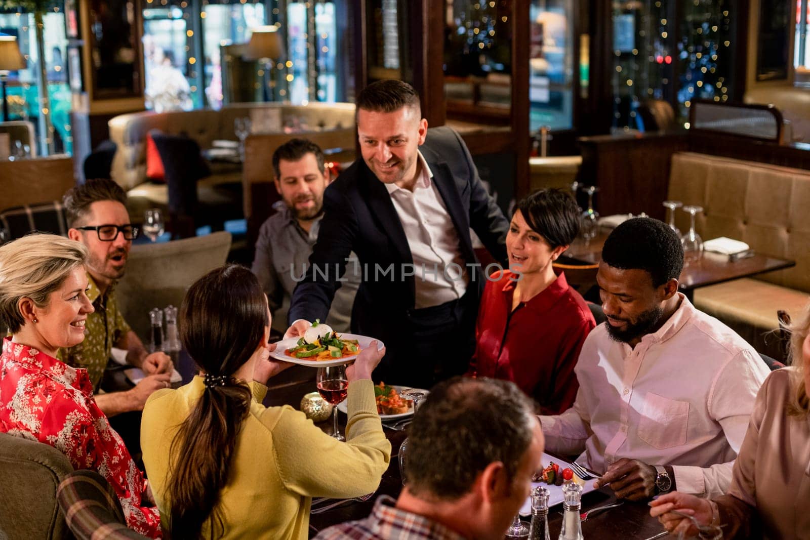 Mature waiter passing a plate of food over to a woman who is sitting with a small group of friends.