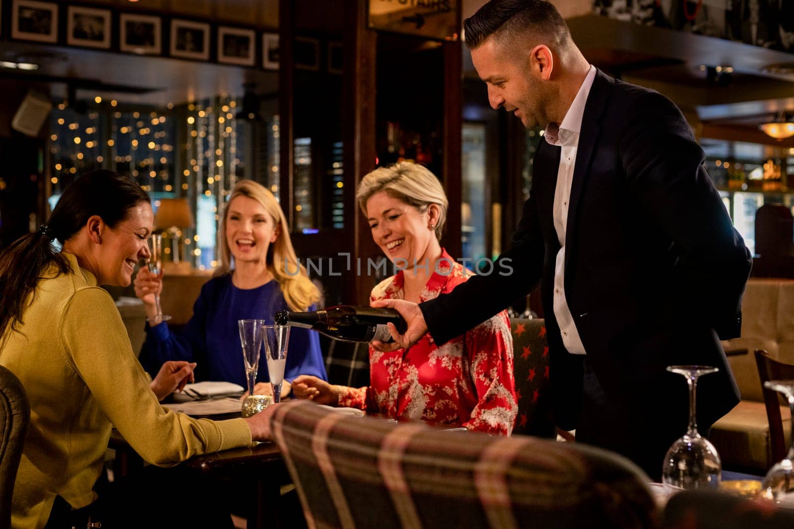 Mature waiter pouring a glass of prosecco for a table with three mature women sitting, laughing and smiling.