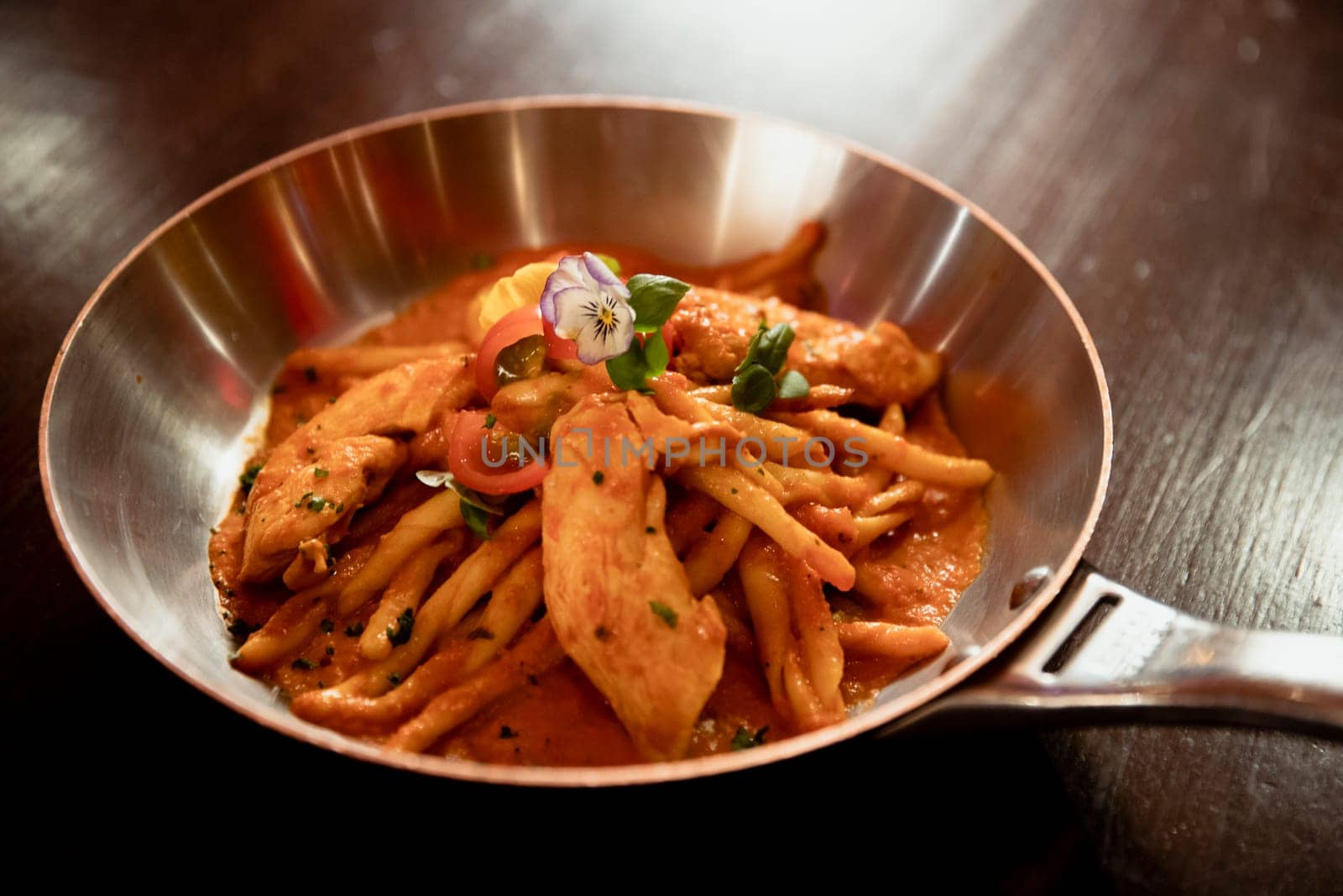 Restaurant style chicken arribiata pasta presented in a silver frying pan on a wooden table in a restaurant.
