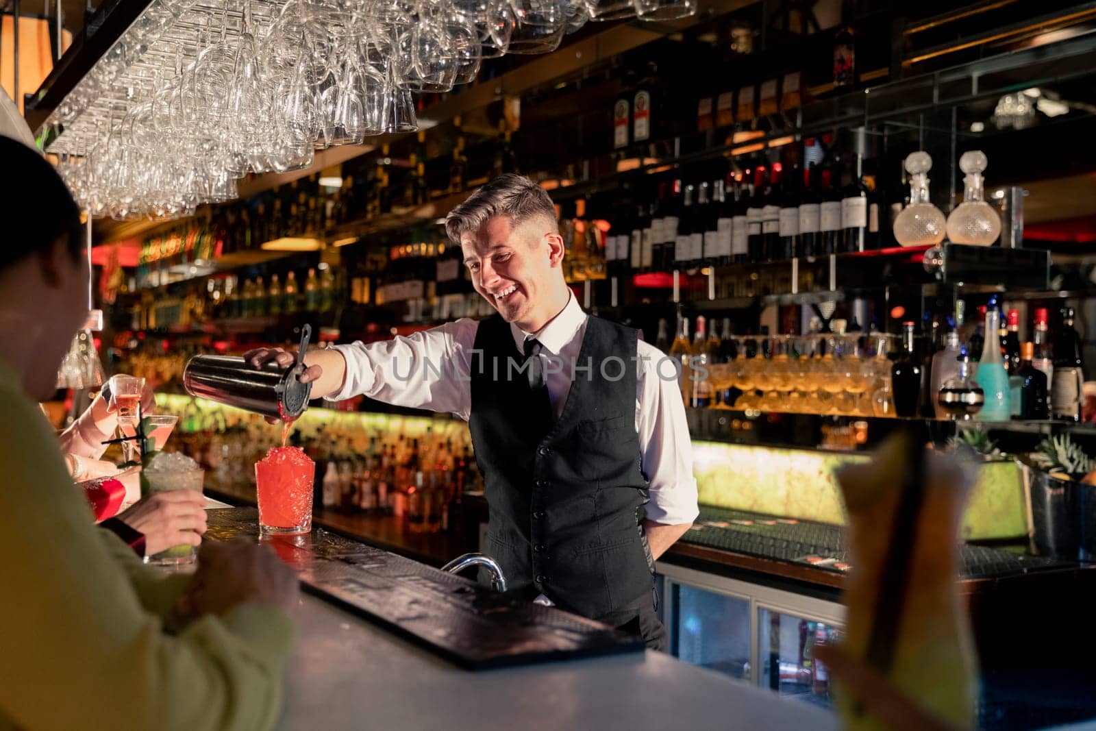 Young bartender pouring a cocktail out of a coktail shaker at the bar counter.