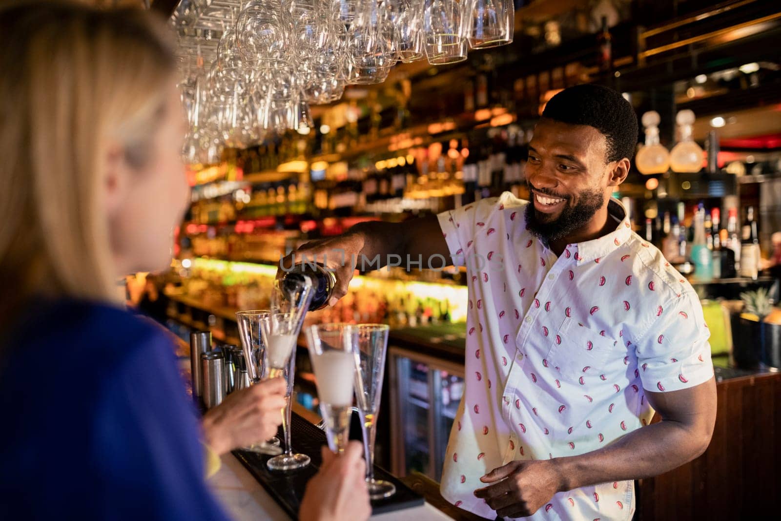 Bartender filling up a mature womans glass with prosecco. She is sitting at the bar counter with her friends.