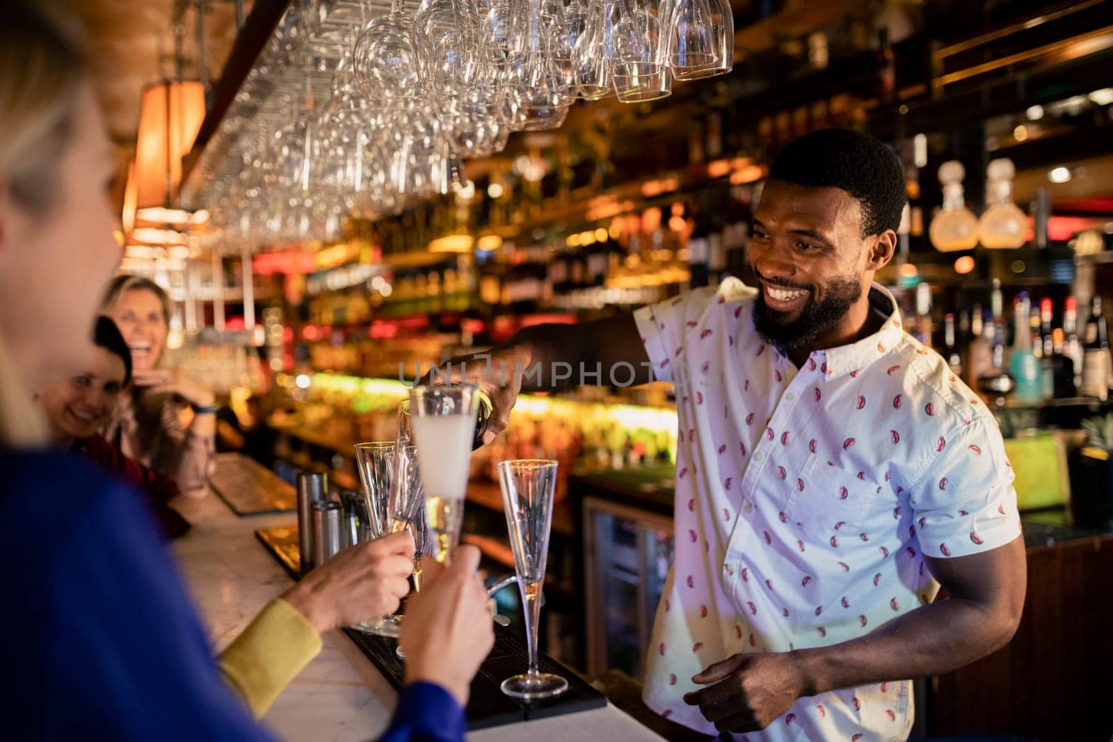Bartender filling up a mature womans glass with prosecco. She is sitting at the bar counter with her friends.