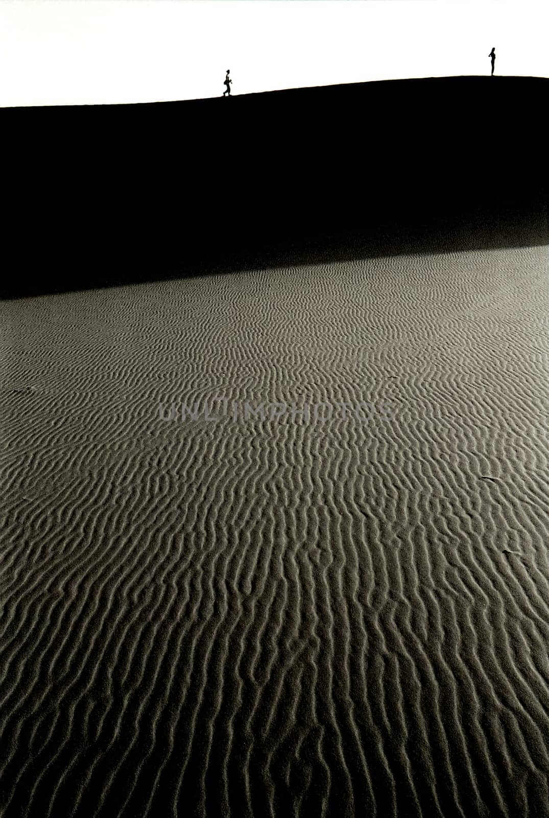 Black and white view of two young women standing high on sand dune of the desert, the first is in contemplation while the other is approaching her walking, Merzouga, Morocco