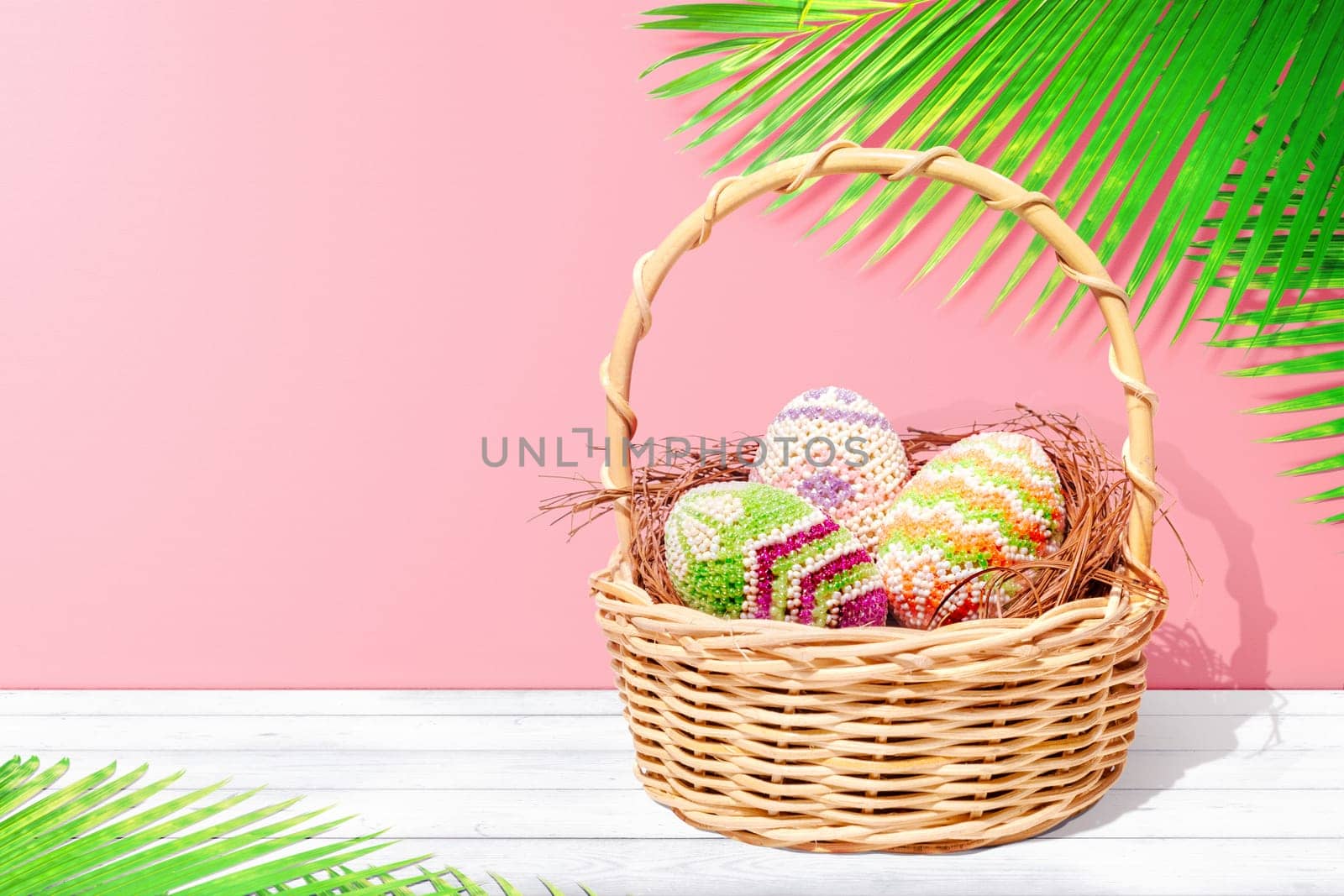 Colorful easter eggs on the nest in wooden basket on white wooden table with palm leaves and pink wall background. Happy Easter