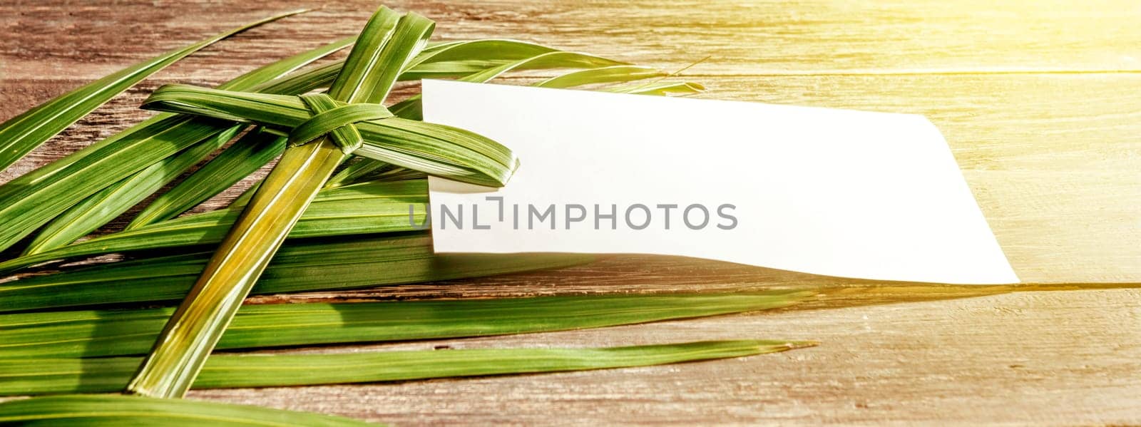 Closeup view of cross shape of palm leaf and palm branches with white blank paper and ray in wooden background. Palm Sunday concept