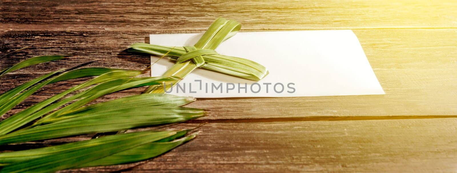 Closeup view of cross shape of palm leaf and palm branches with white blank paper and ray in wooden background. Palm Sunday concept