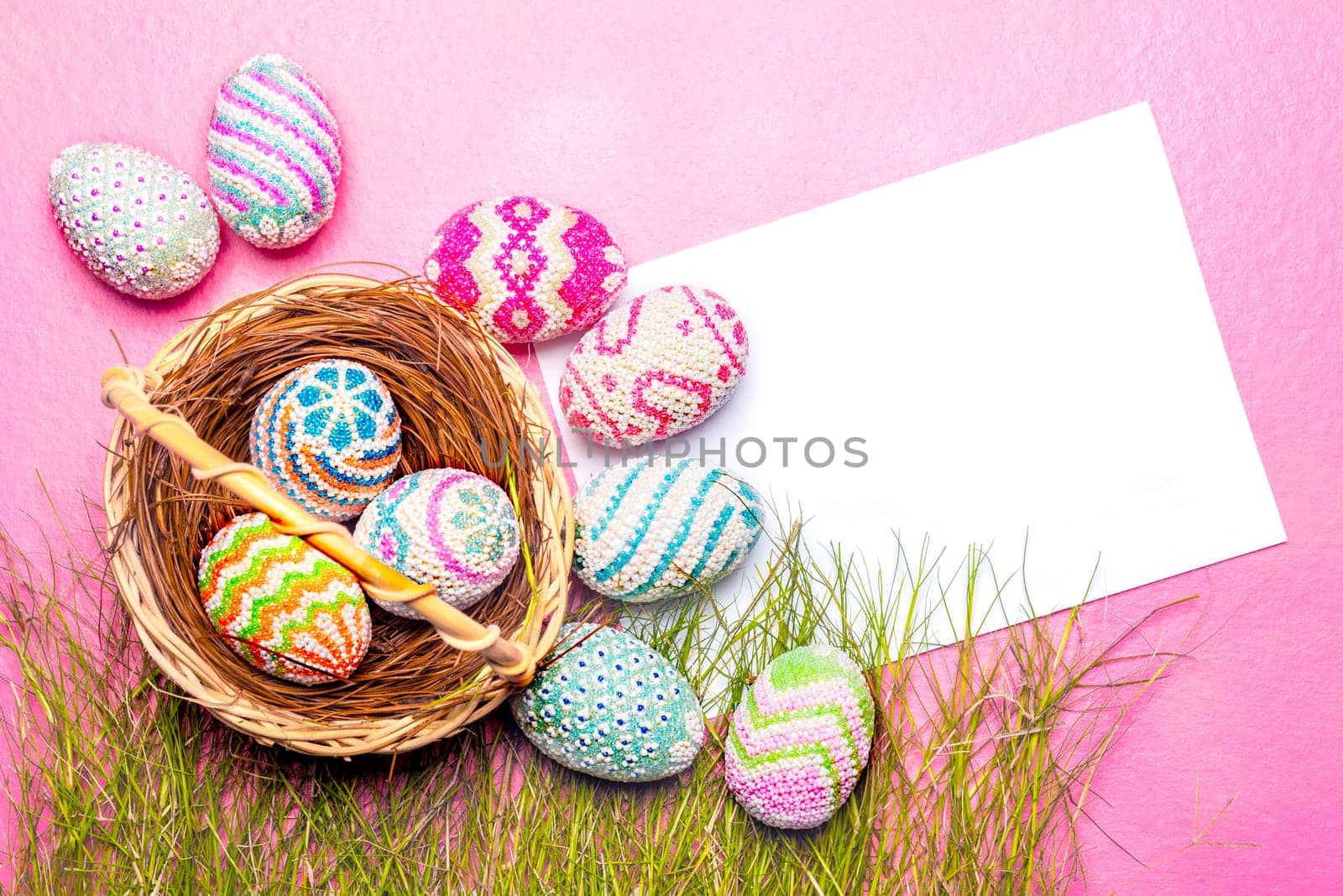 Colorful easter eggs in the nest and grass with blank white paper on the pink background. Happy Easter