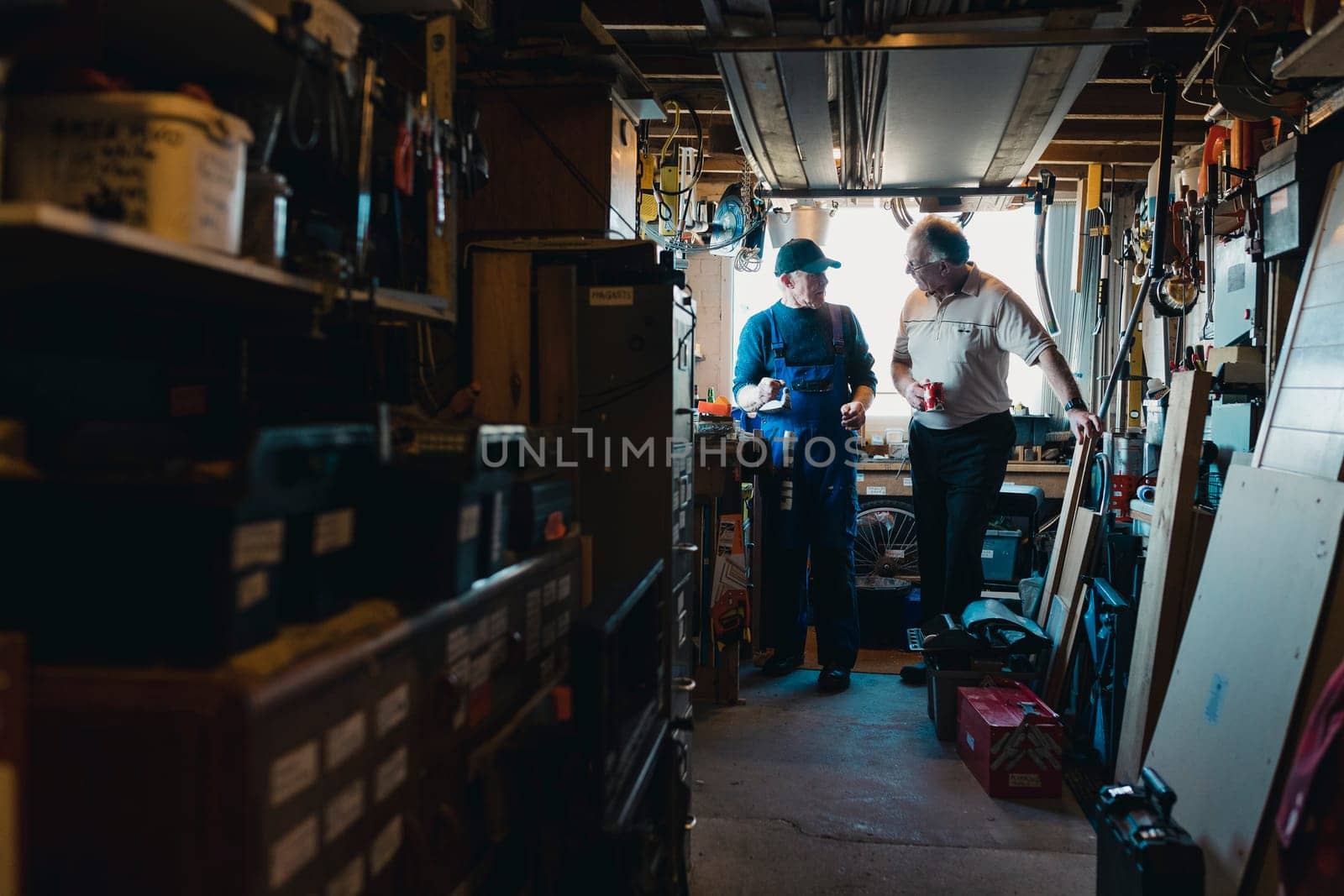 Two senior men are in a construction workshop. One man is explaining something and the other man is drinking a cup of tea while listening.