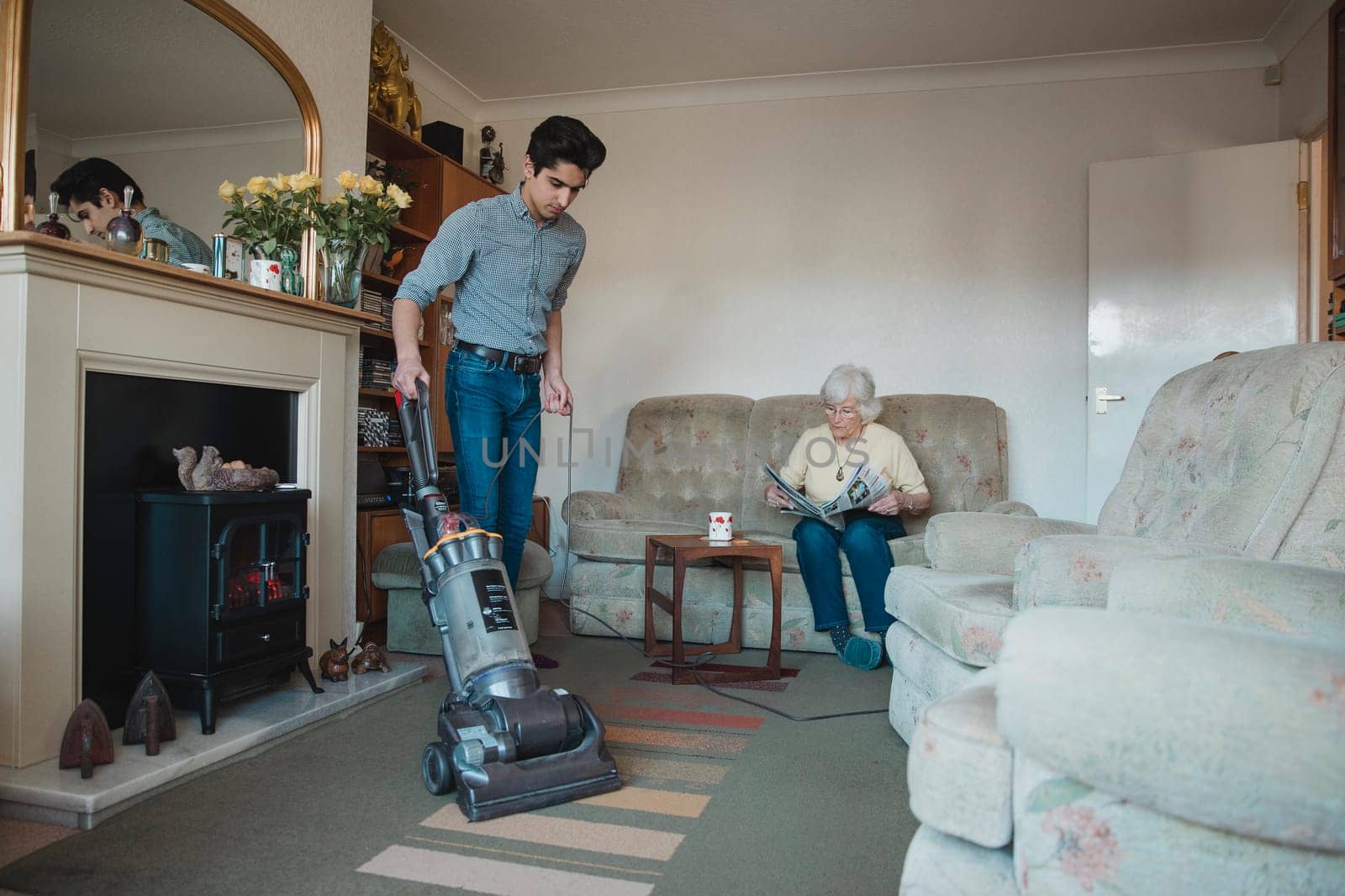 Teenage boy is hoovering his grandmother's living room for her while she reads a newspaper.