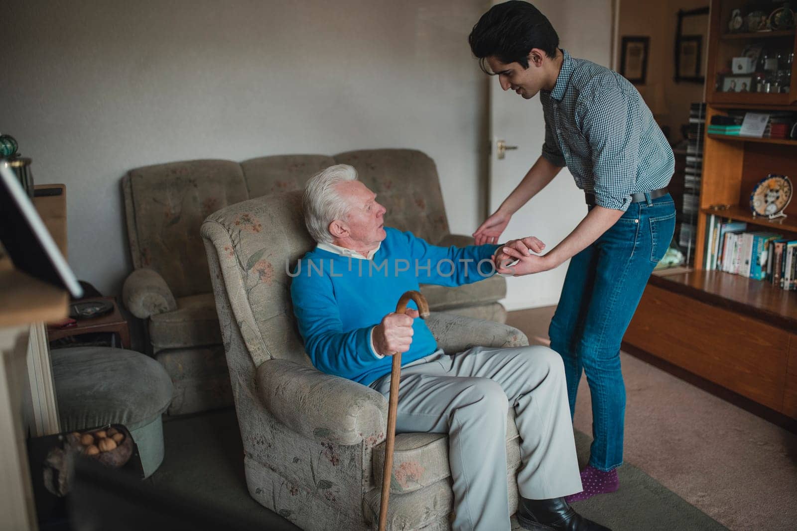 Teenage boy is helping his grandfather out of his armchair.