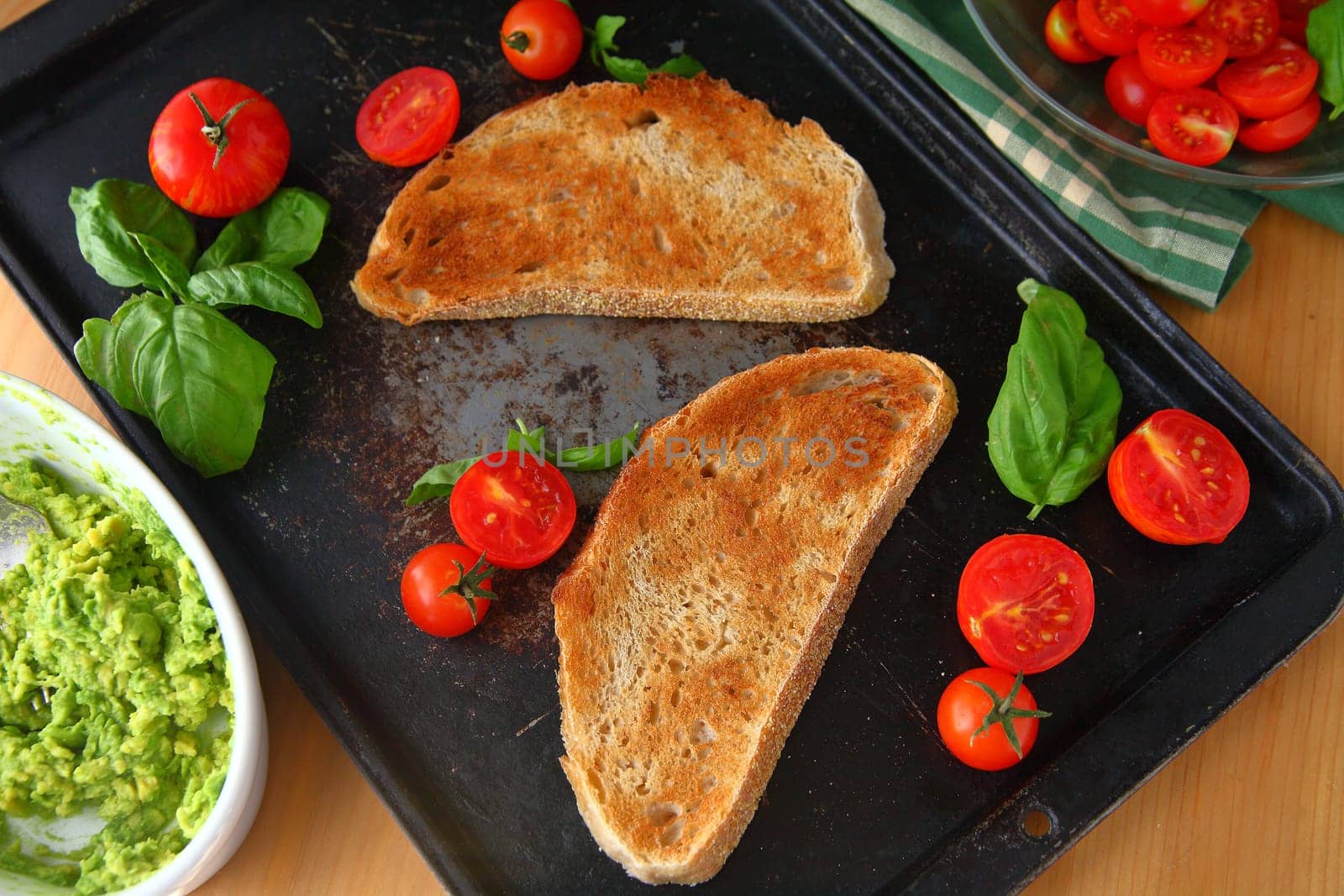 Overhead view of toast slices, mashed avocado, cherry tomatoes and basil on old baking pan