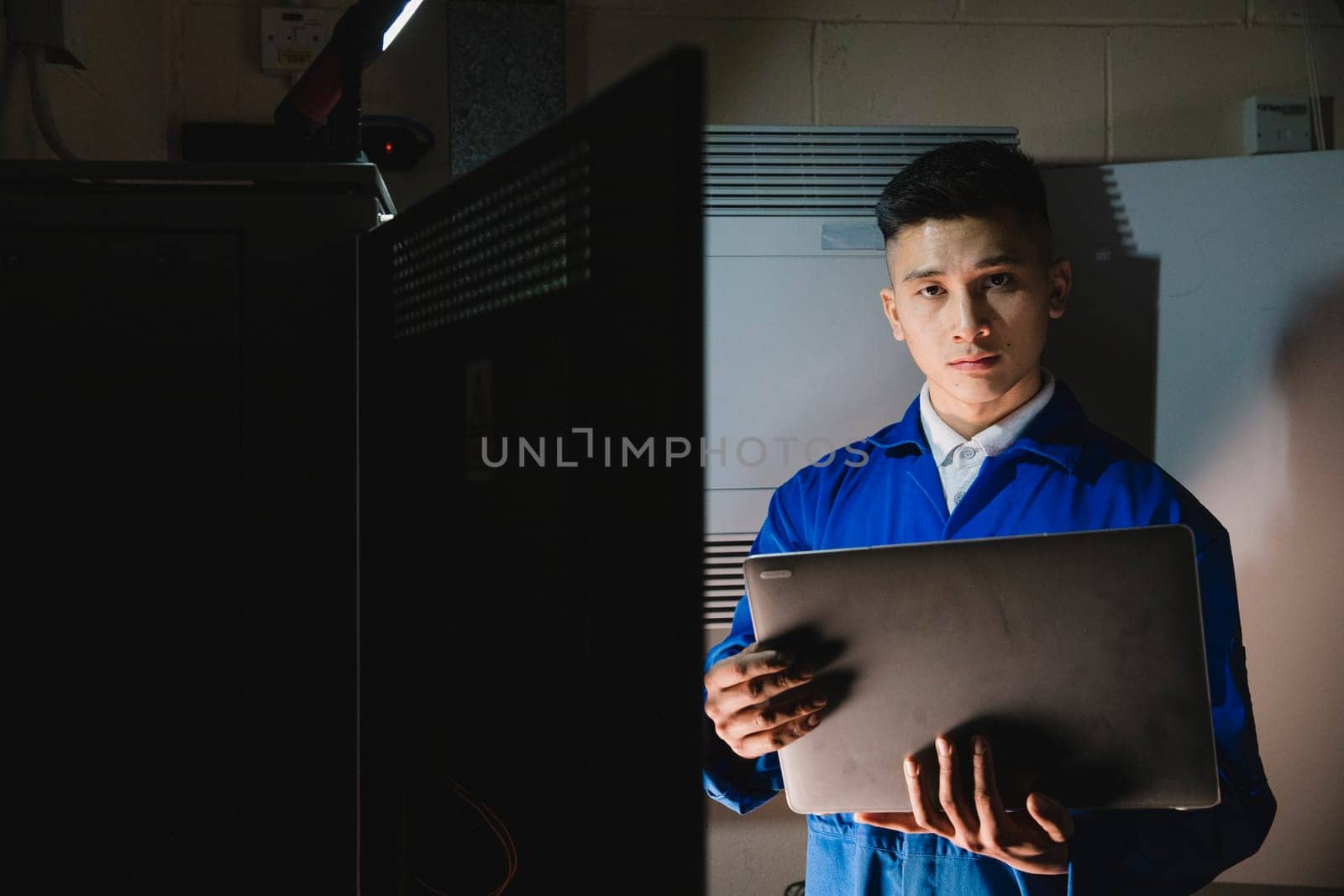 Young man is looking at the camera while in the server room with a laptop.
