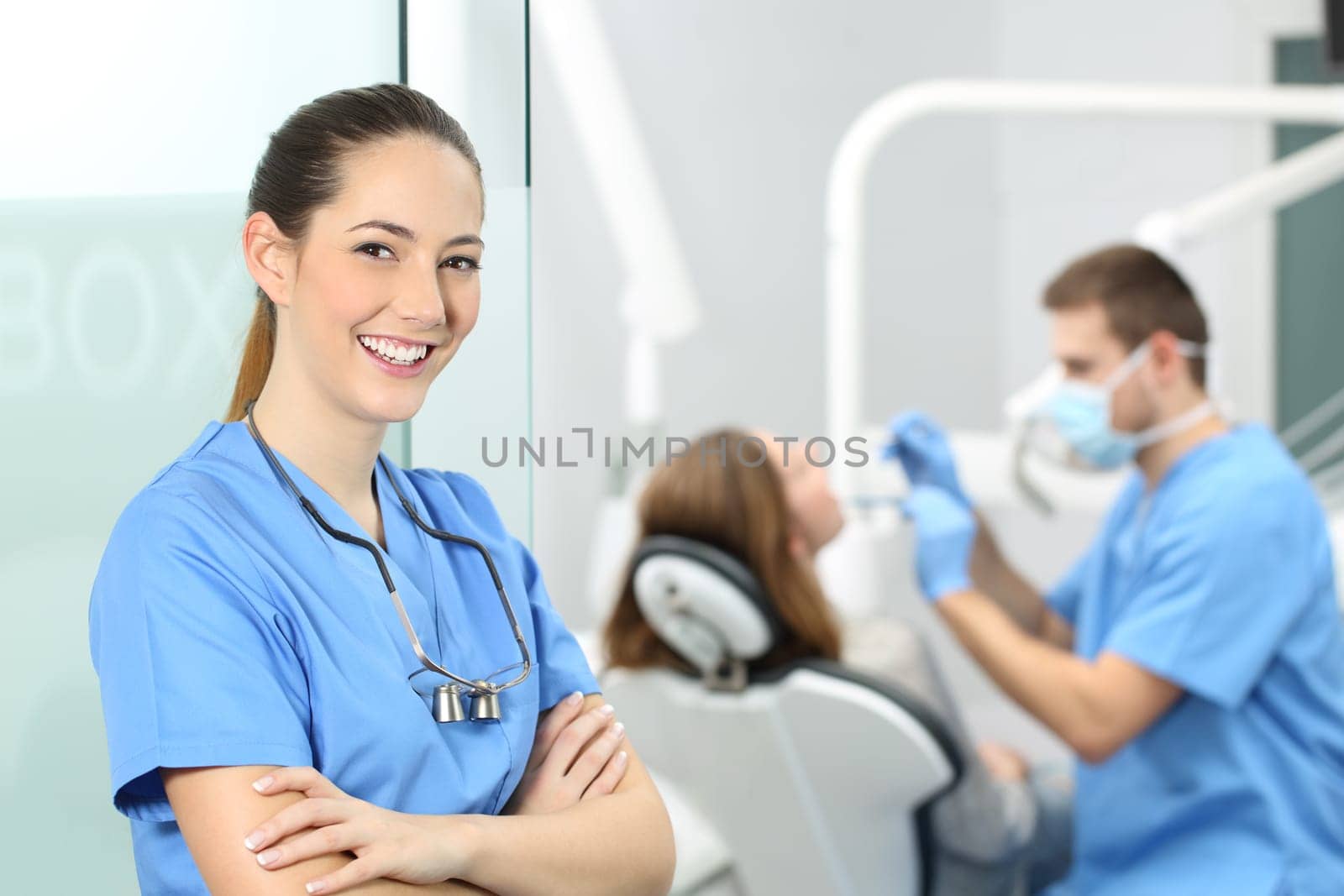 Dentist female with crossed arms wearing blue coat posing and looking at you at consultation with a doctor working with a patient in the background