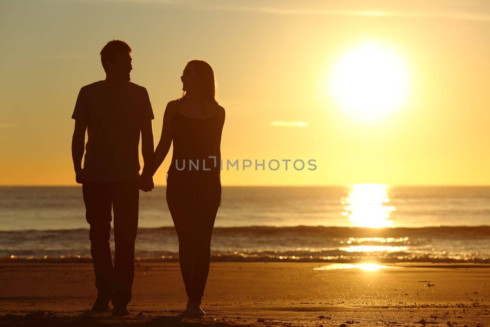 Front view of a full body of a couple silhouette walking together on the beach at sunset in summer