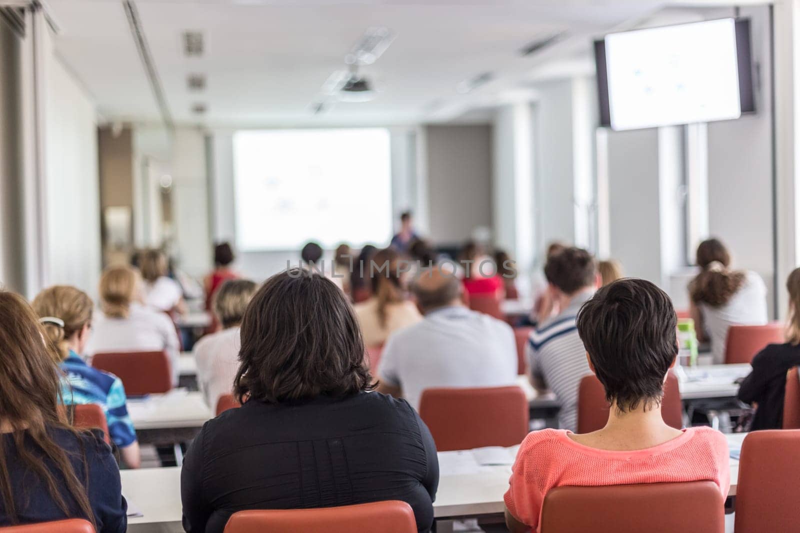 Female speaker giving presentation in lecture hall at university workshop. Audience in conference hall. Rear view of unrecognized participant in audience. Scientific conference event.