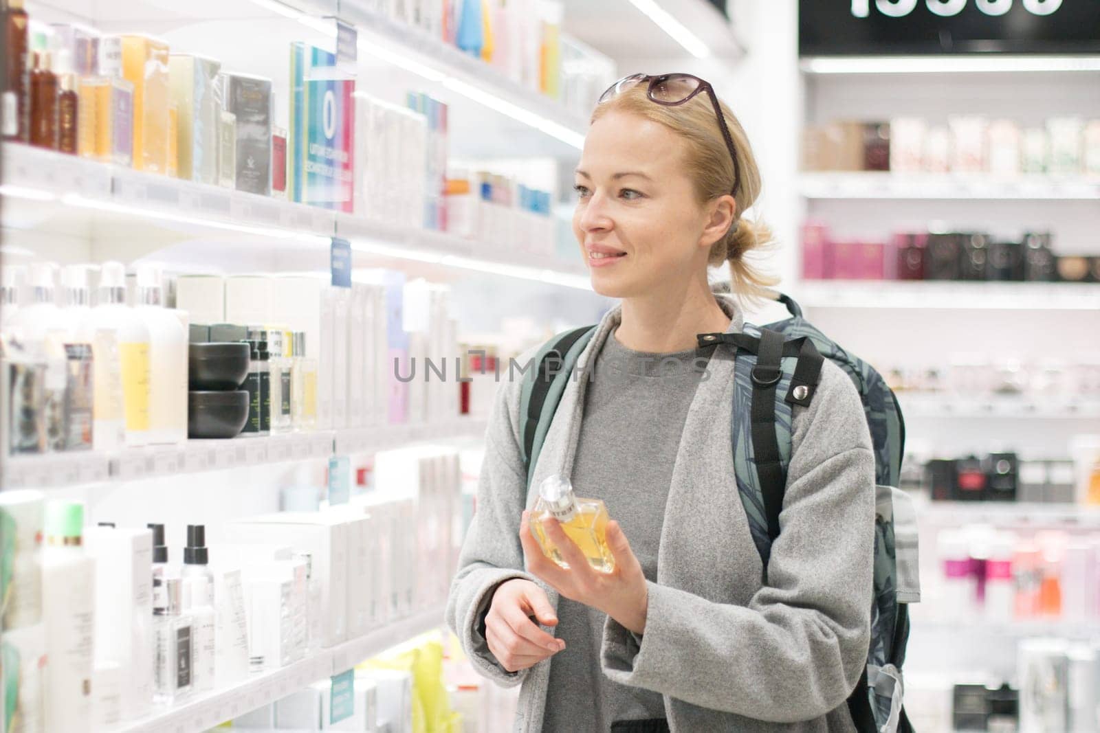 Blond young female traveler wearing coat and travel backpack choosing perfume in airport duty free store. Casual lady testing and buying cosmetics on the go in a beauty store.