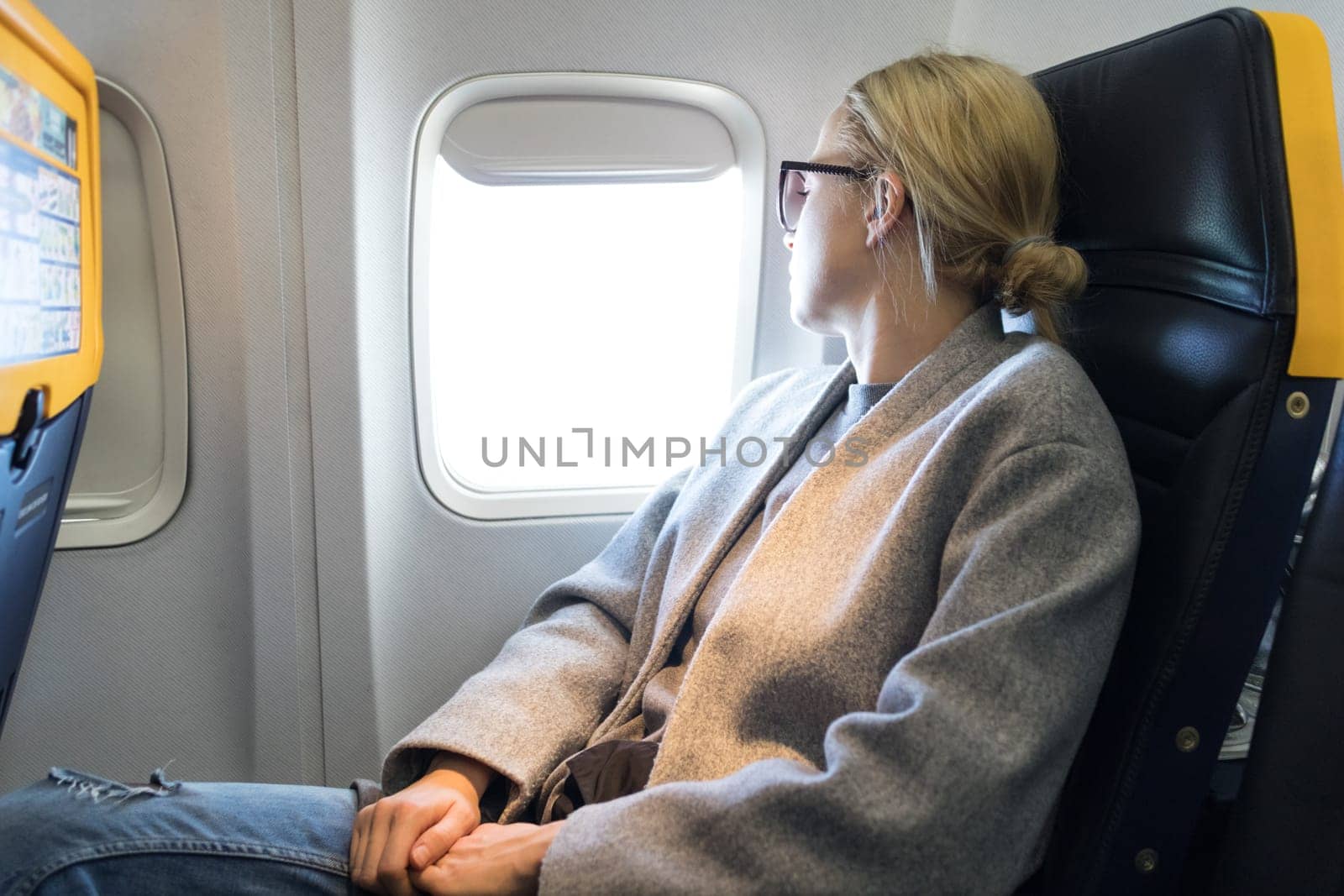Thoughtful blonde casual caucasian woman wearing glasses, looking through the window while traveling by airplane. Commercial transportation by planes.