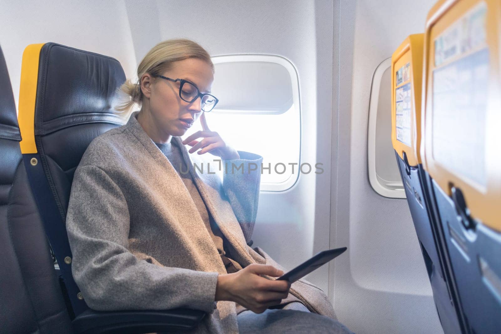Blonde casual caucasian woman wearing glasses reading on digital e-reader while traveling by airplane. Commercial transportation by planes.