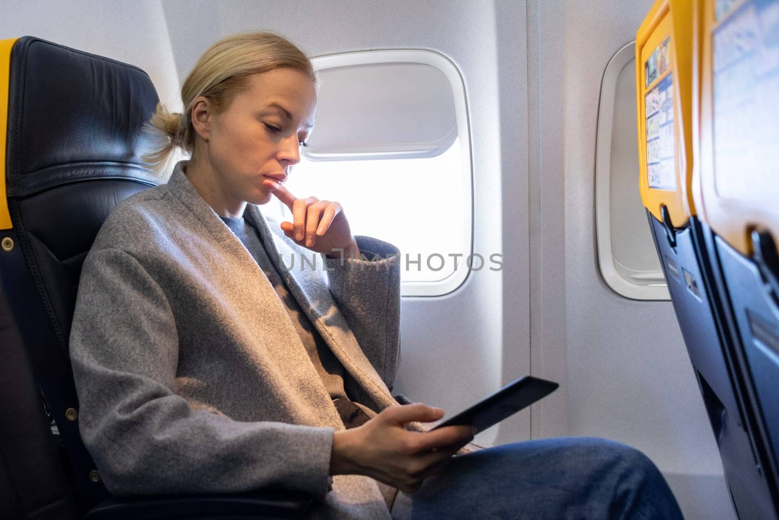 Thoughtful blonde casual caucasian woman reading e-book on digital e-reader while traveling by airplane. Commercial transportation by planes.