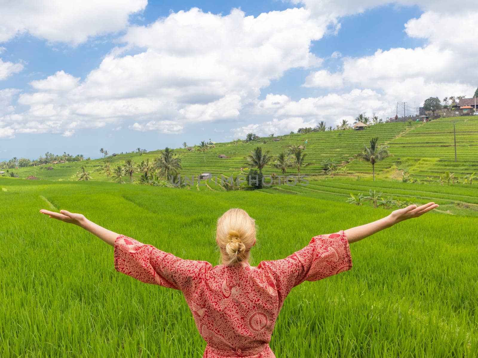 Relaxed caucasian woman wearing red asian style kimono, arms rised, enjoying life of beautiful green rice fields on Bali, feeling free, relaxed and happy. Concept of well being, conected to nature,...