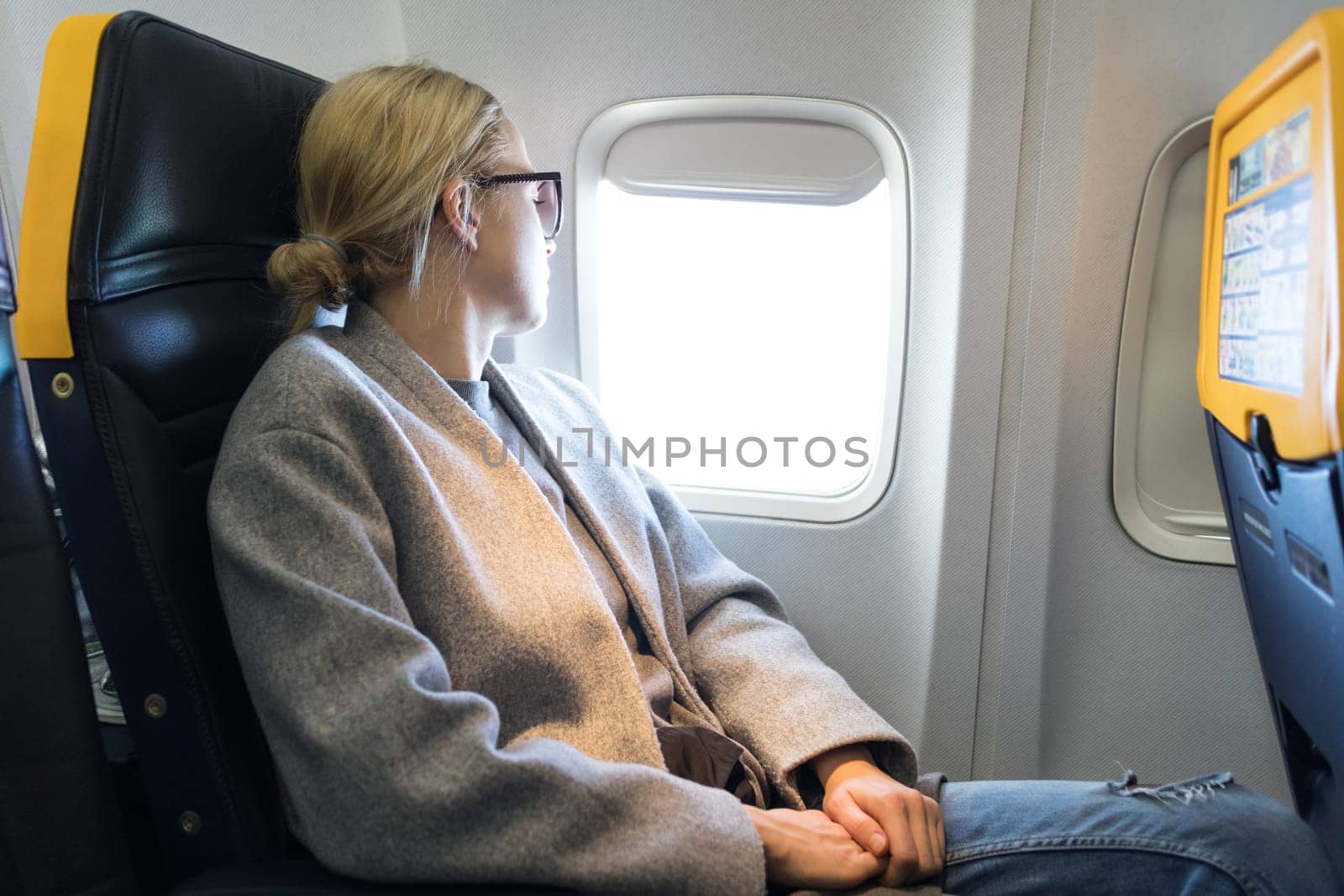 Thoughtful blonde casual caucasian woman wearing glasses, looking through the window while traveling by airplane. Commercial transportation by planes.