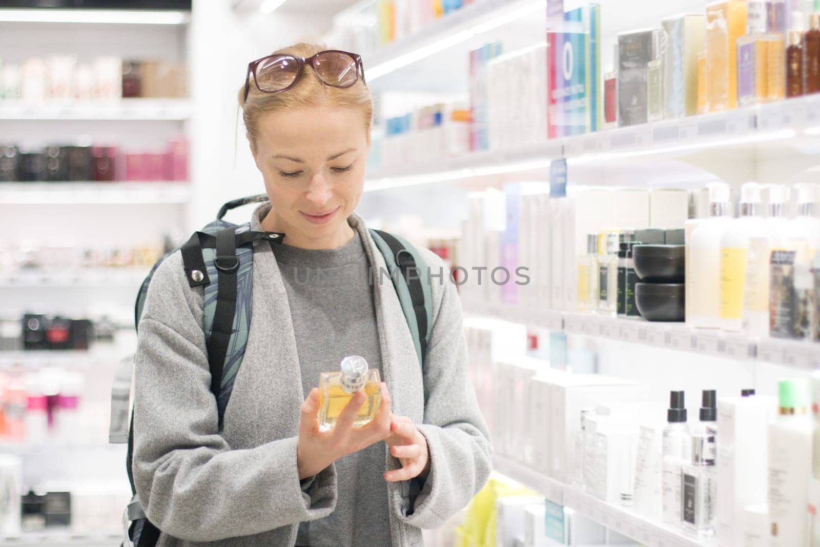Blond young female traveler wearing coat and travel backpack choosing perfume in airport duty free store. Casual lady testing and buying cosmetics on the go in a beauty store.