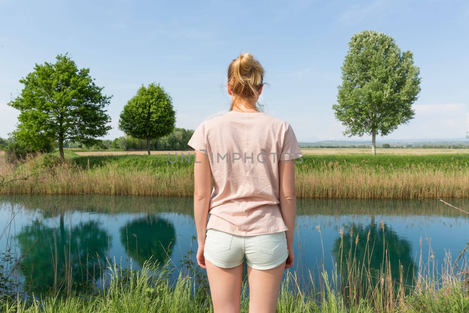 Young woman wearing casual summer clothes enjoying beautiful view of peaceful countryside. Trees on the meadow reflecting in the river.