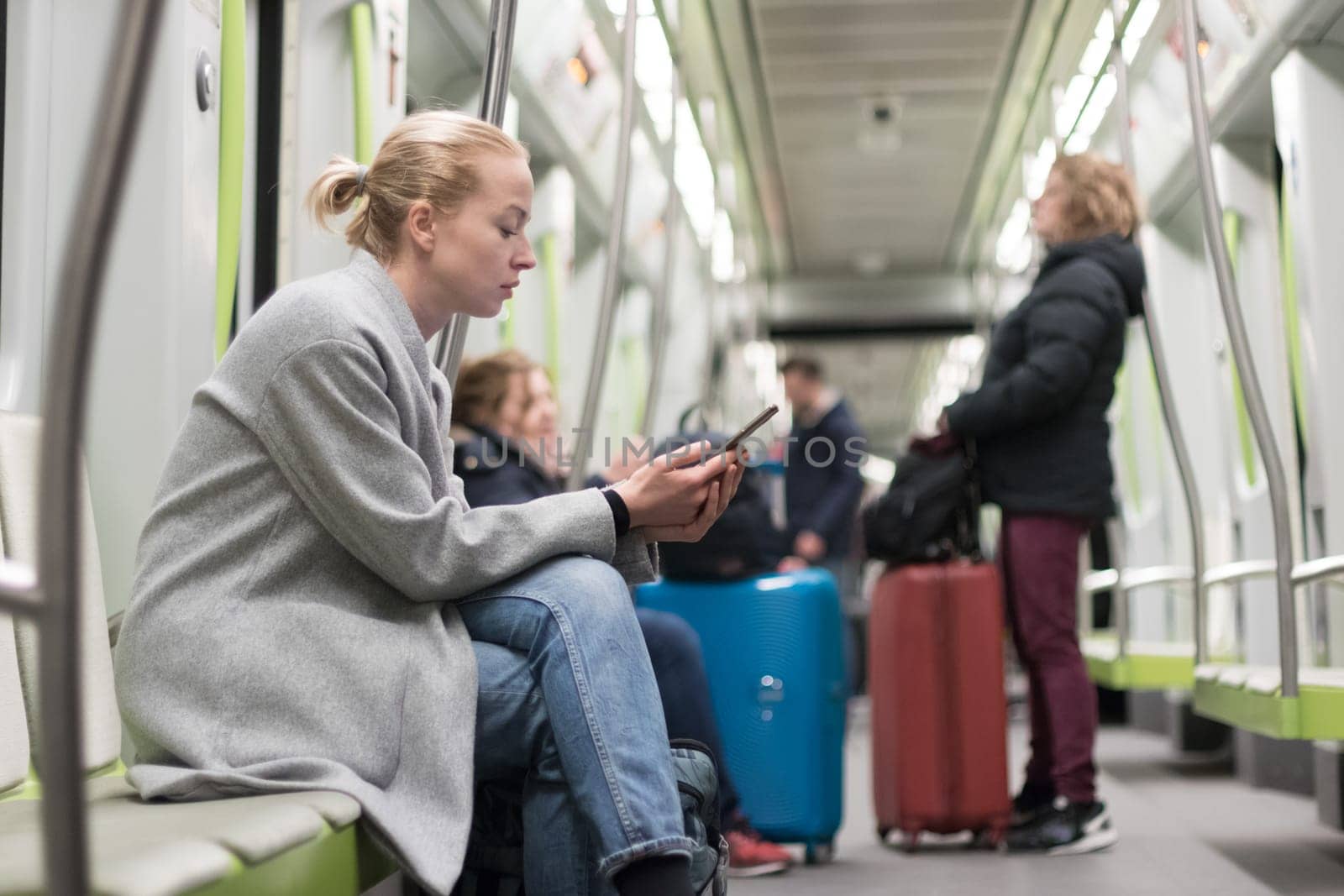 Beautiful blonde caucasian woman wearing winter coat reading on the phone while traveling by metro. Public transportation concept.