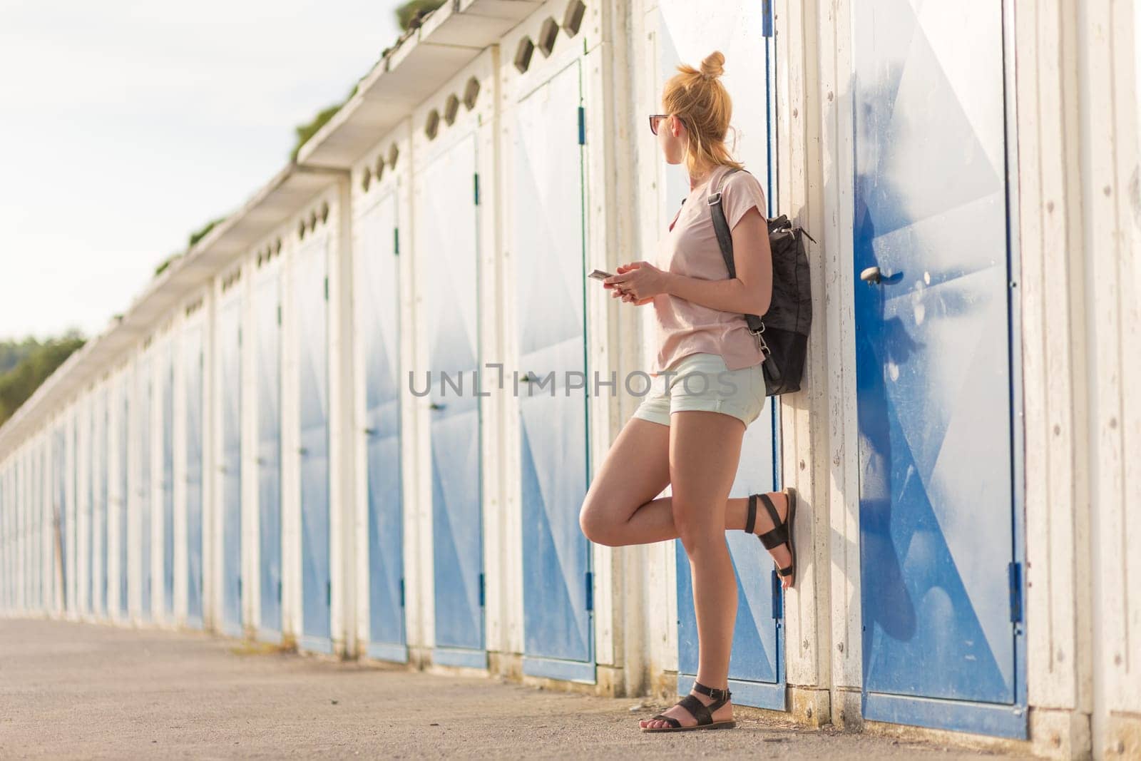 Blonde young female traveler wearing summer style clothing, holding mobile phone, against retro blue beach dressing rooms at summer time vacation in Sistiana, Italy.
