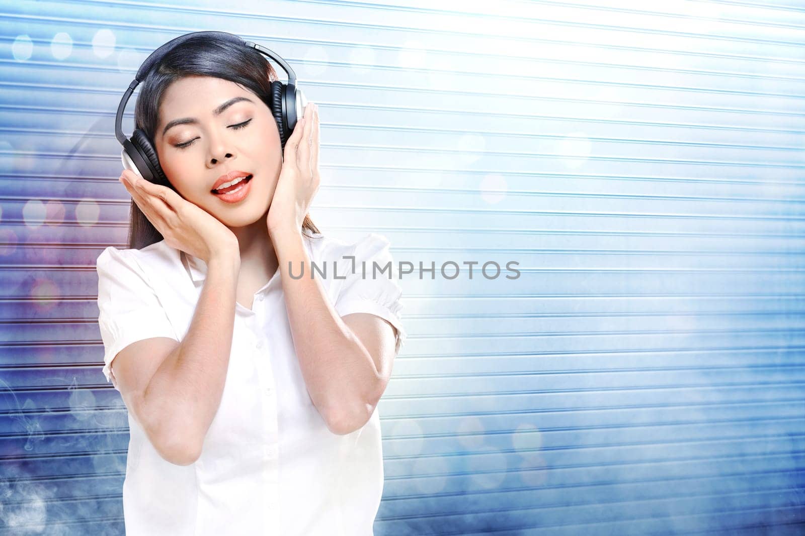 Pretty asian woman in white shirt listening music with headphones over white roller shutter door with smoke and colorful light reflection background