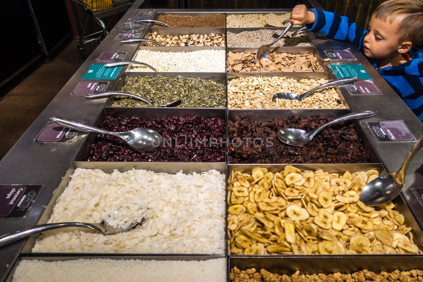 London, England - July 2018 : Young boy reaching out with a ladle to try the dehydrated fruits including raisins and pieces of coconut and banana on sale in the market stall