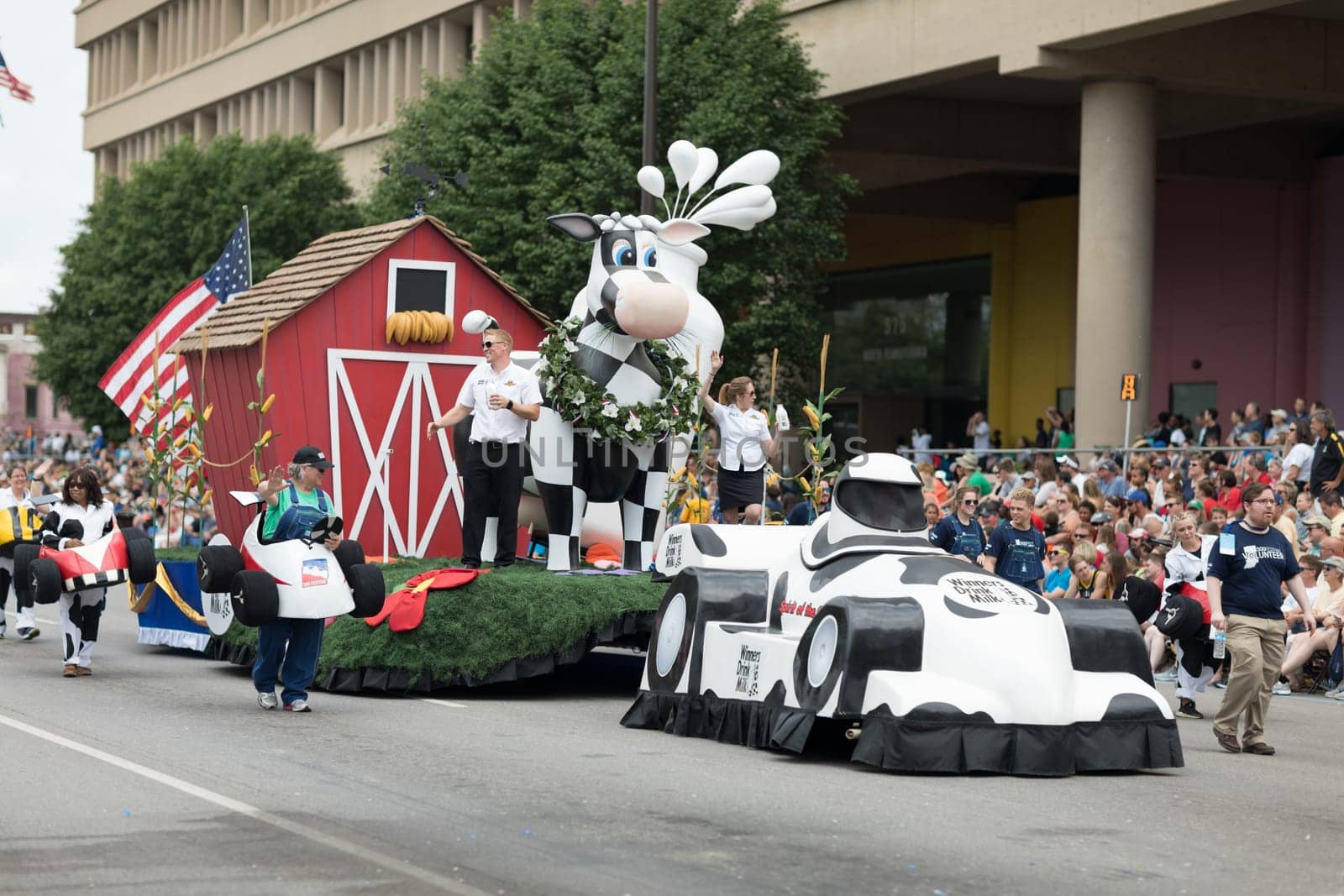 Indianapolis, Indiana, USA - May 26, 2018,  People with race car outfits walking along a float with a giant cow, promoting milk drinking, at the Indy 500 Parade