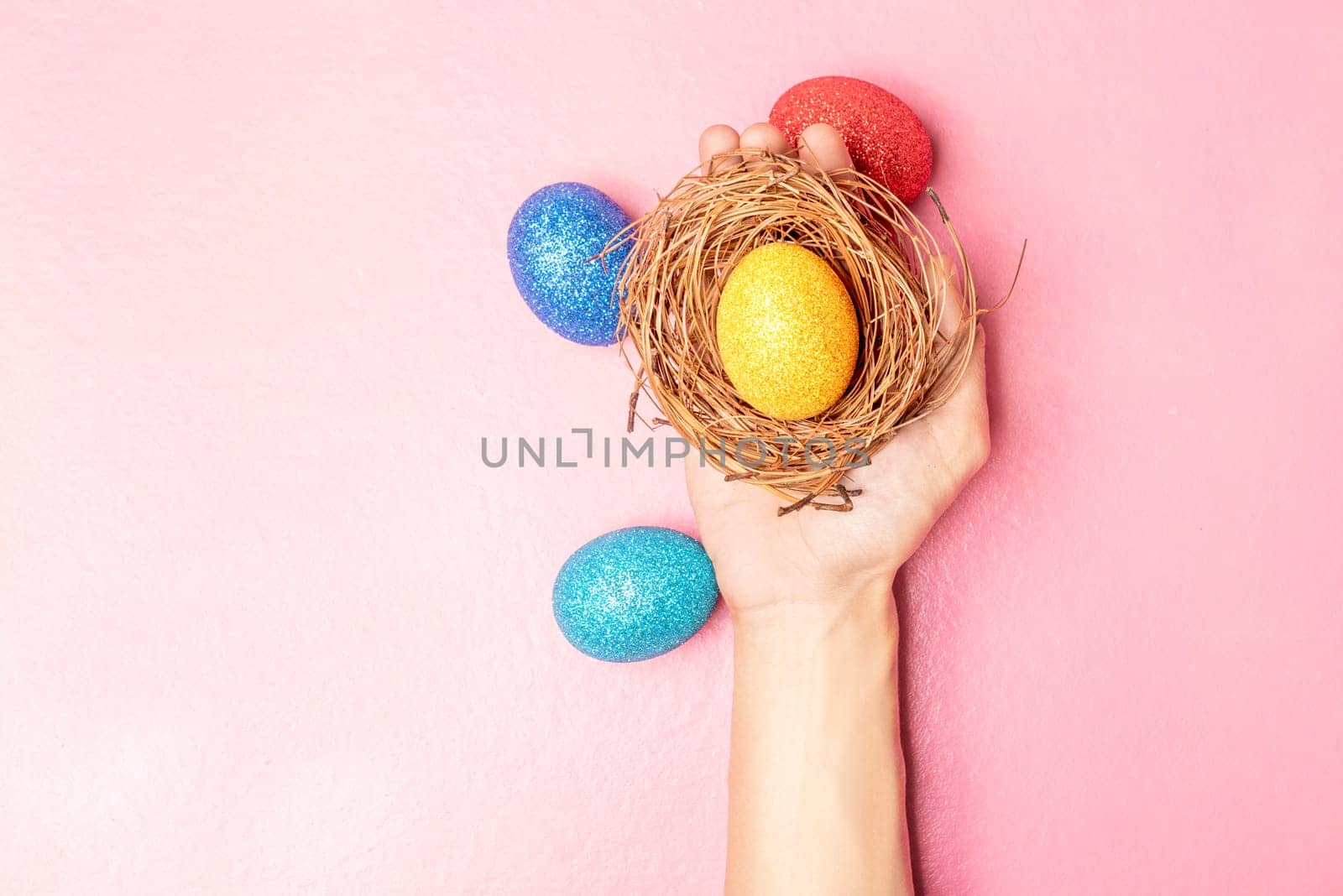 Human hand holding the nest with yellow easter egg and colorful easter eggs on pink color background. Happy Easter