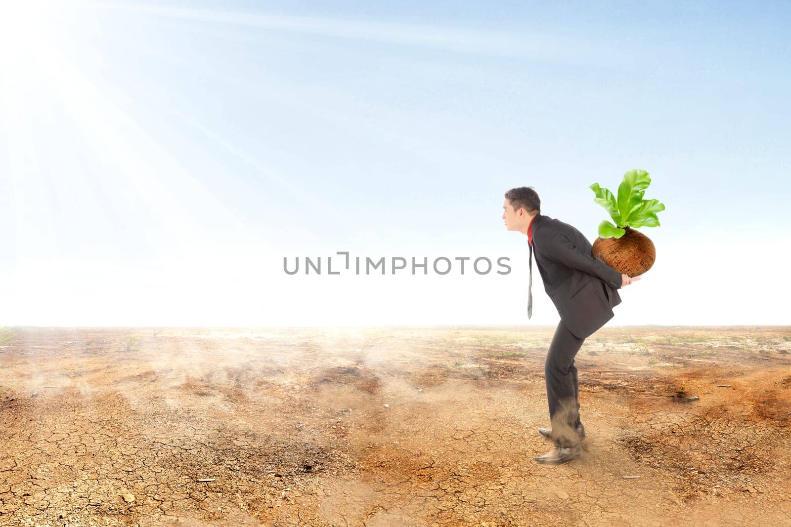 Young asian businessman carrying young plant with green leaves in the pot on the cracked field and sunlight over blue sky background. Earth day concept