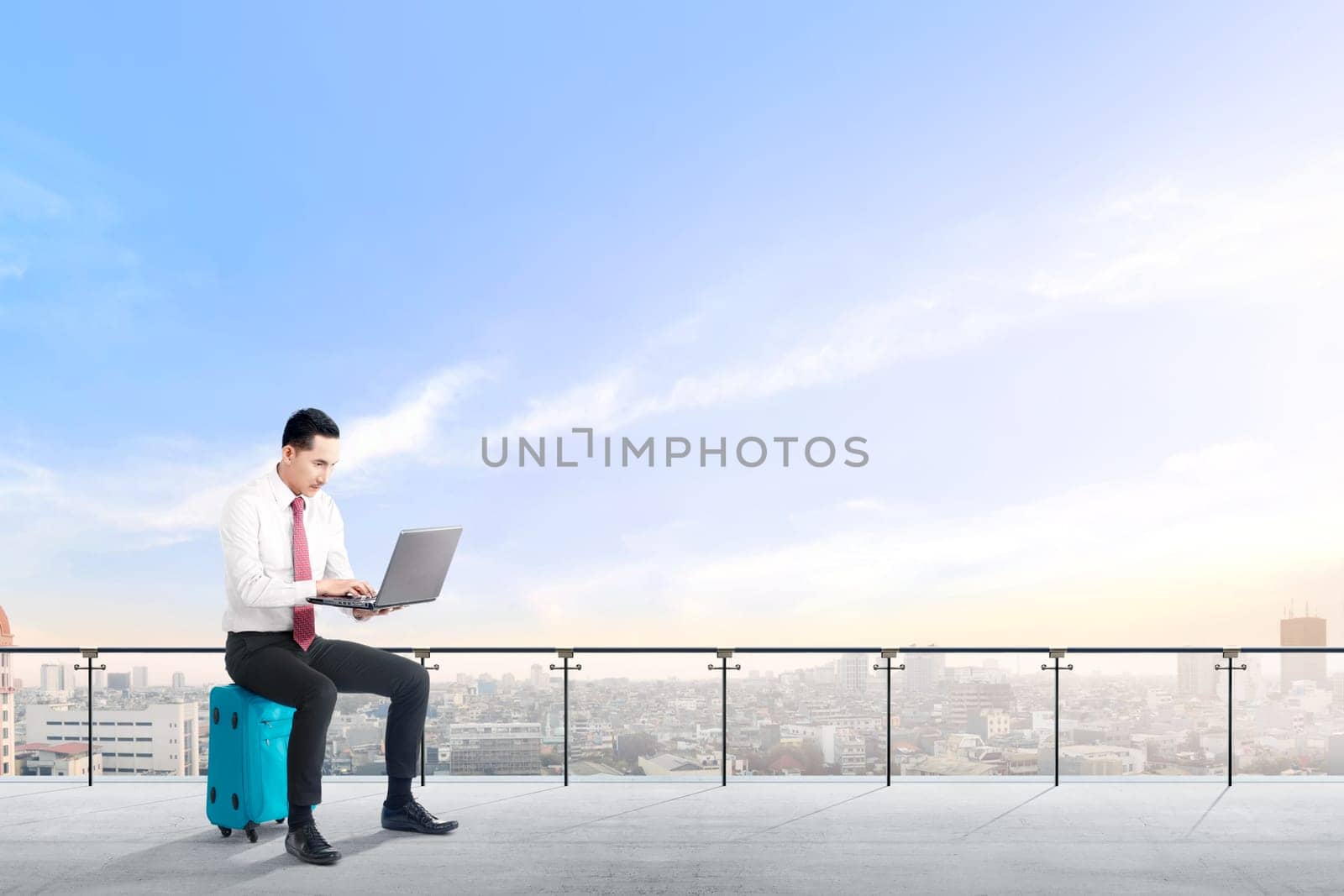 Handsome asian businessman sitting on blue suitcase hold the laptop and working on modern terrace with city view and blue sky background