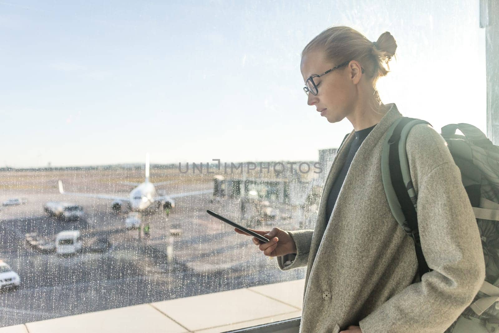 Casually dressed female traveler at airport looking at smart phone device in front of airport gate windows overlooking planes on airport runway.