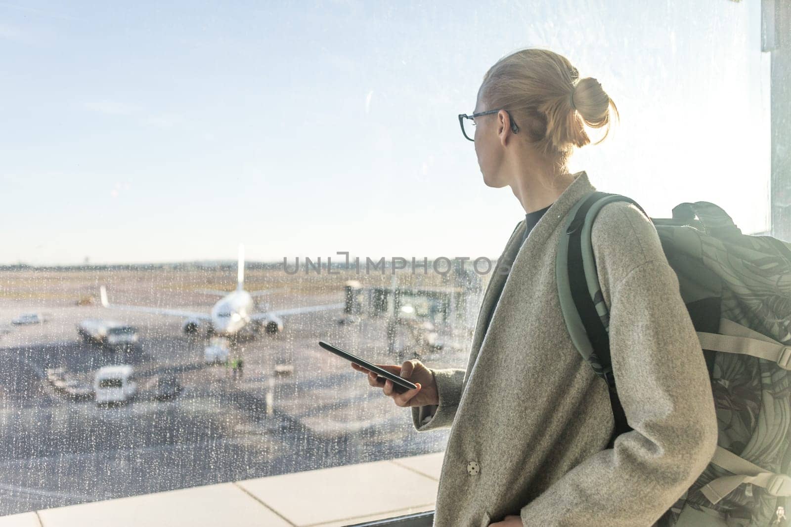 Casually dressed female traveler at airport, holding smart phone device, looking through the airport gate windows at planes on airport runway.