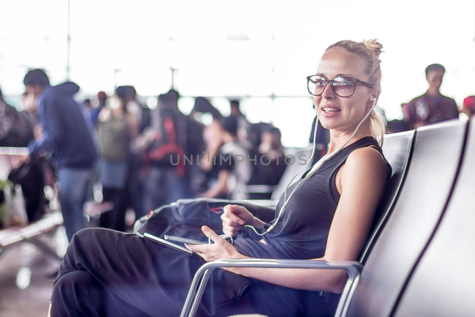 Casual sporty young blond female traveler using her cell phone while waiting to board a plane at the departure gates at the asian airport terminal.