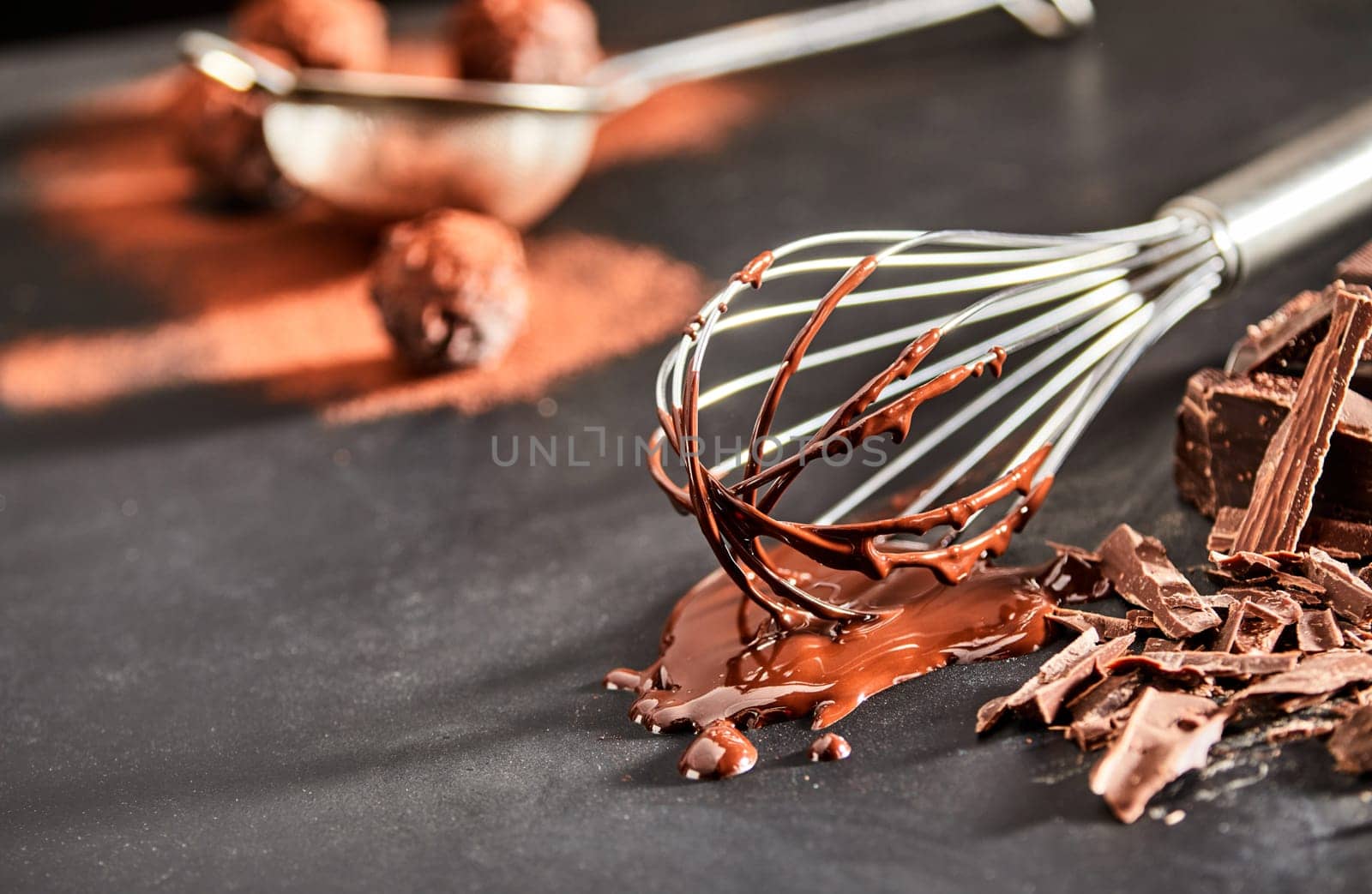 Used metal whisk coated in melted chocolate lying on a kitchen counter with flaked chocolate and copy space