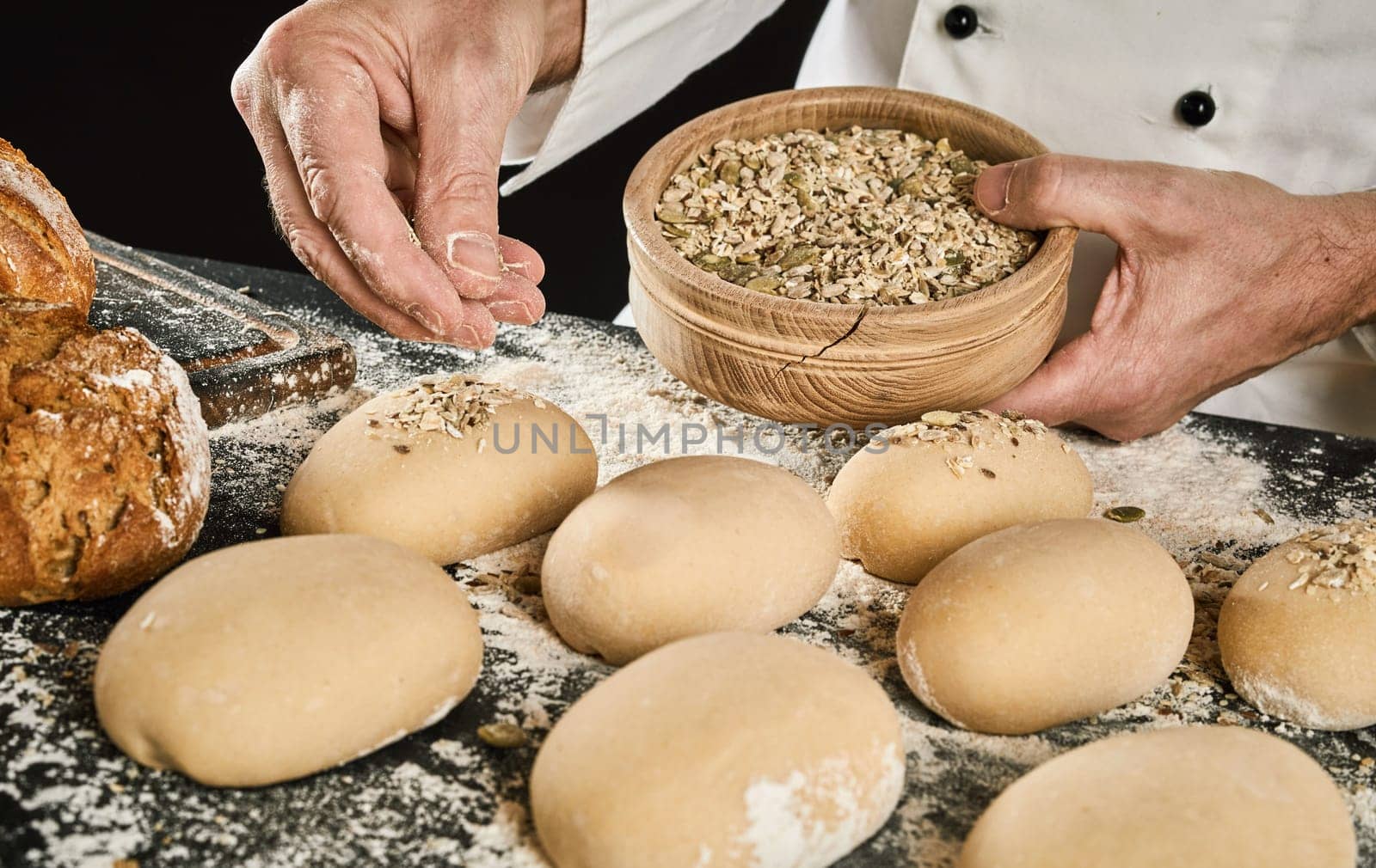 Close-up of baker hands sprinkling seeds on raw bread buns on table from wooden bowl in his hand. Bread preparation process in professional bakery or restaurant