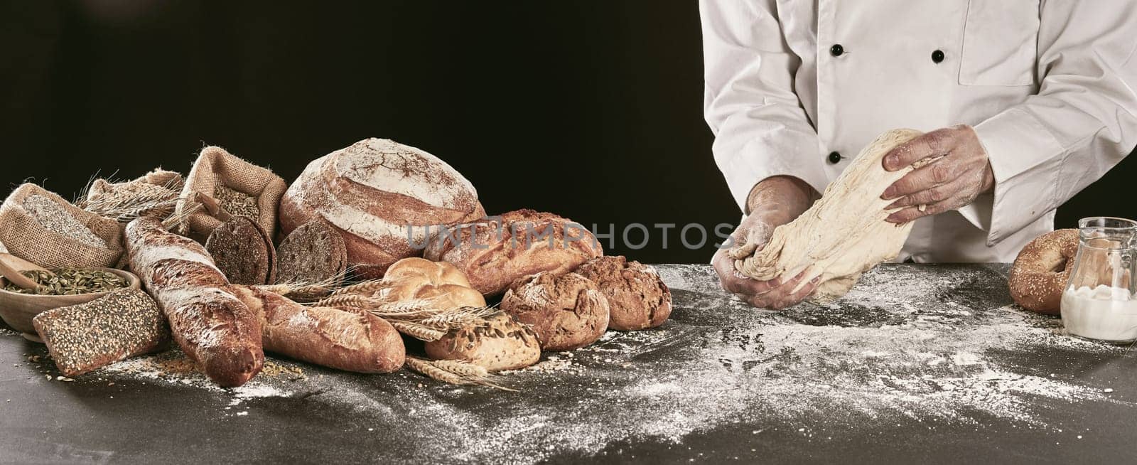 Chef or baker kneading dough while making speciality bread on a black counter with a display of a variety of his products alongside, close up on hands panorama banner