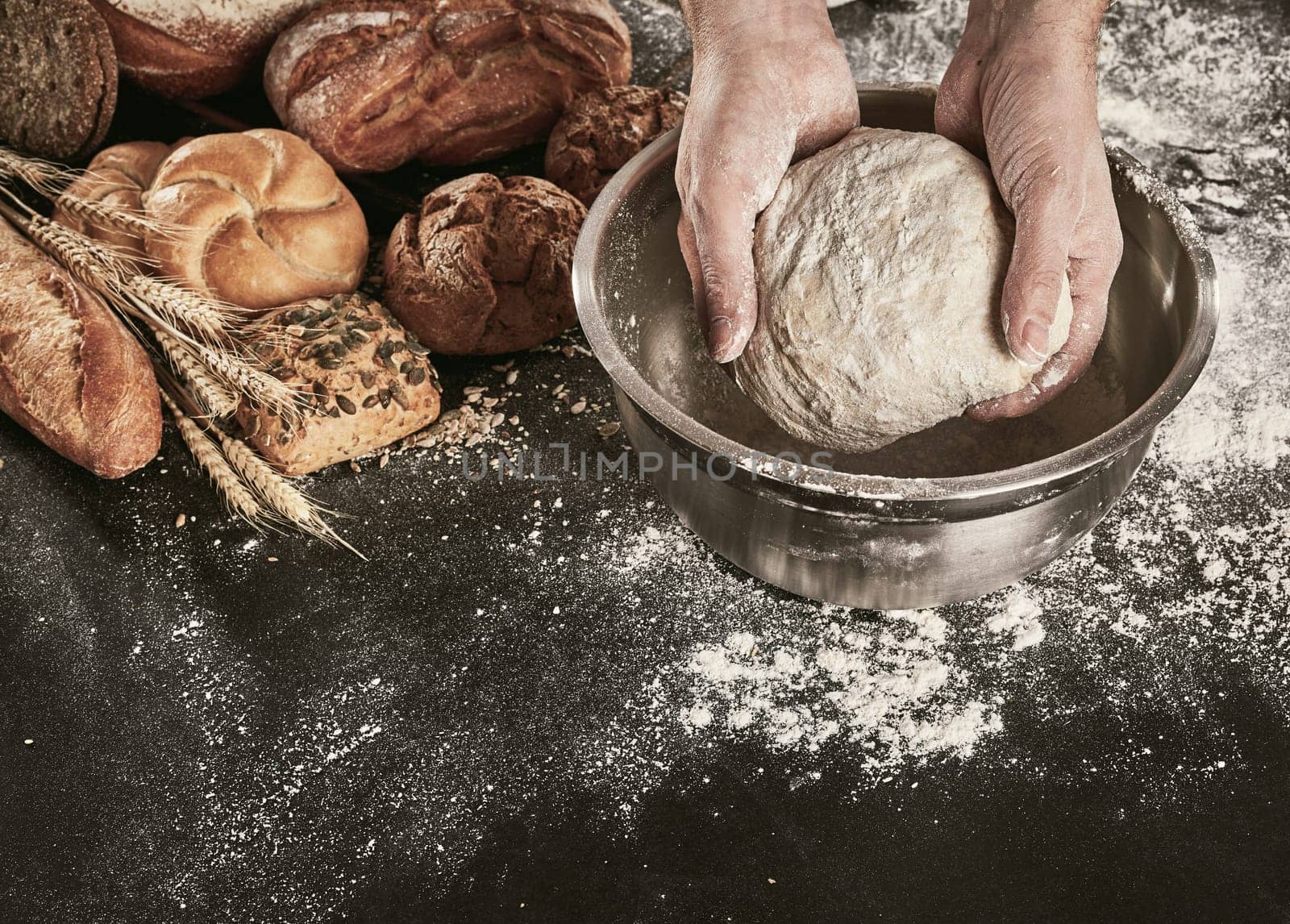 Hands of a baker kneading dough in a metal mixing bowl on a flour covered counter with an assortment of speciality breads alongside