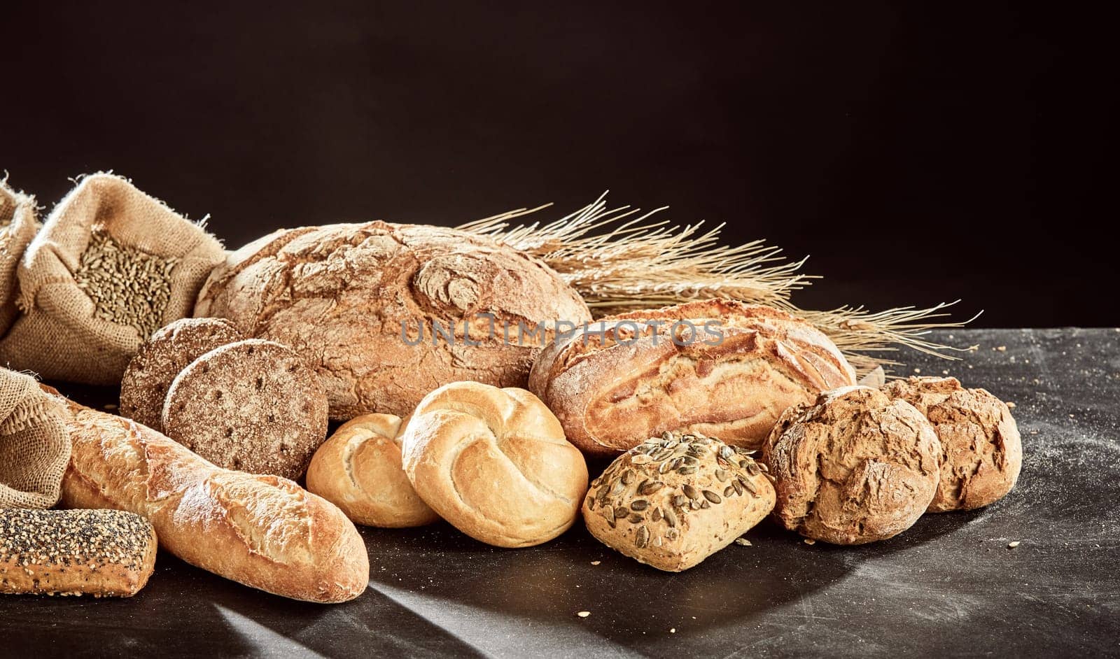 Fresh bread loaves assortment piled on dark table surface with grains of rye and wheat, against black background