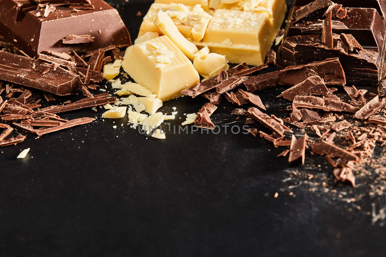 Shavings and pieces of white and milk chocolate, viewed in close-up from high angle on dark table surface background with copy space. Cooking ingredients concept