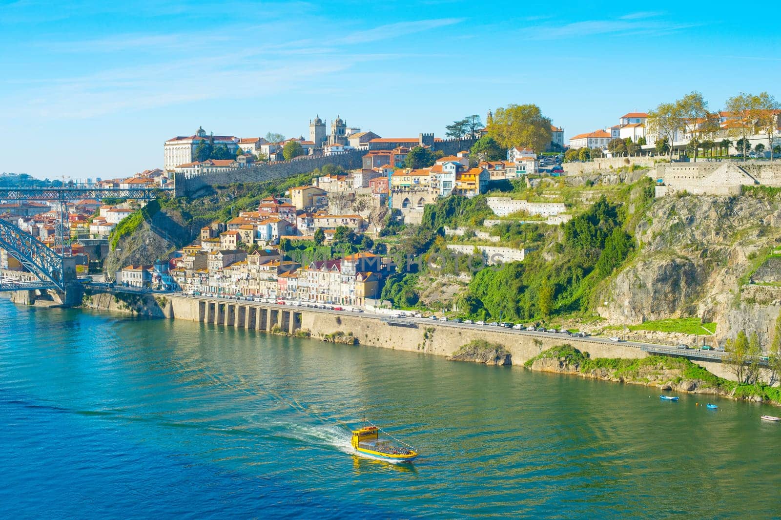 Tourist boat sailing by Douro with Porto cityscape on green riverbank in bright sunlight, Portugal